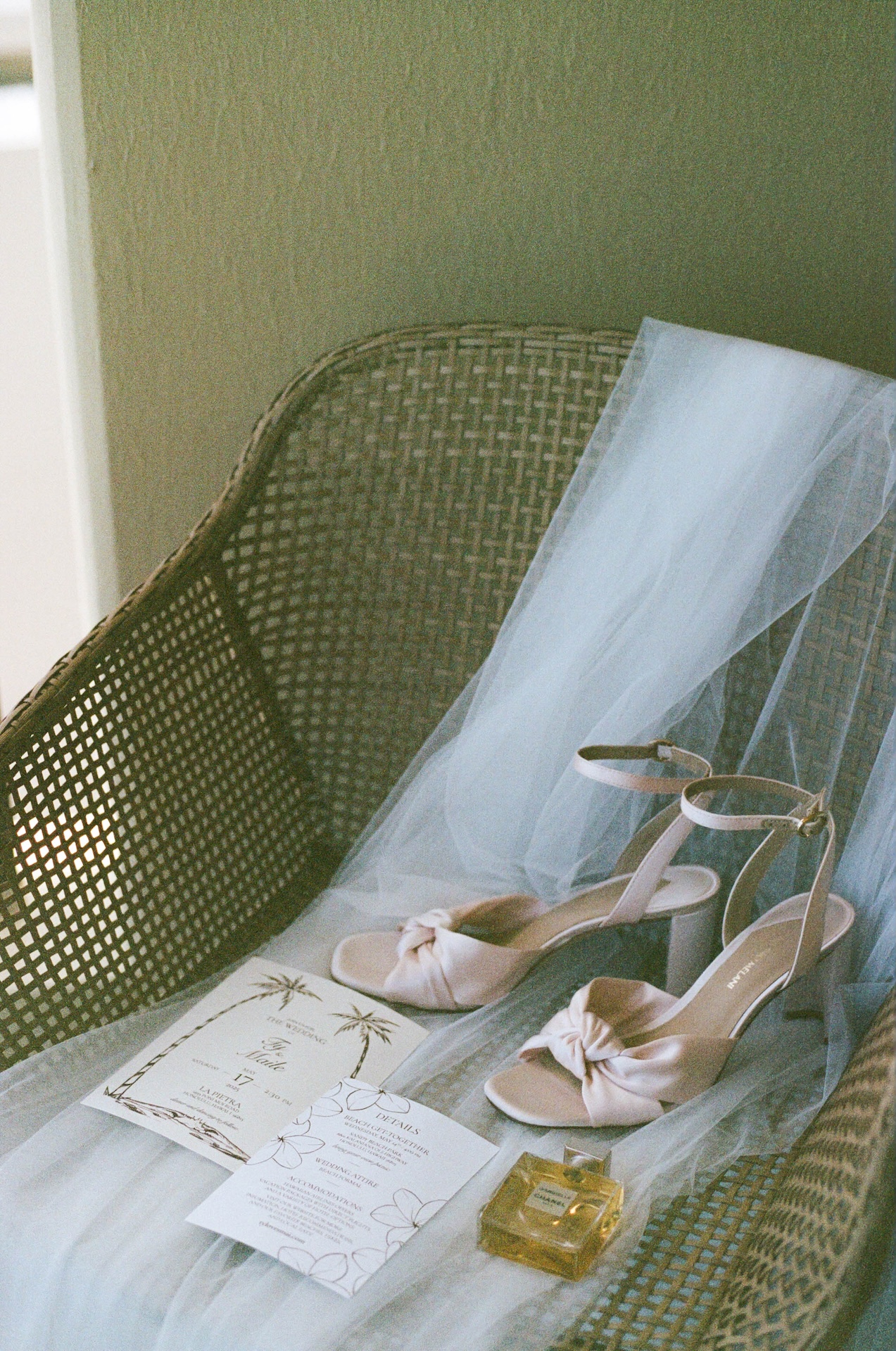 Flat lay of blush wedding heels, a tropical invitation suite, and perfume atop a wicker chair and veil — styled by a destination wedding photographer.
