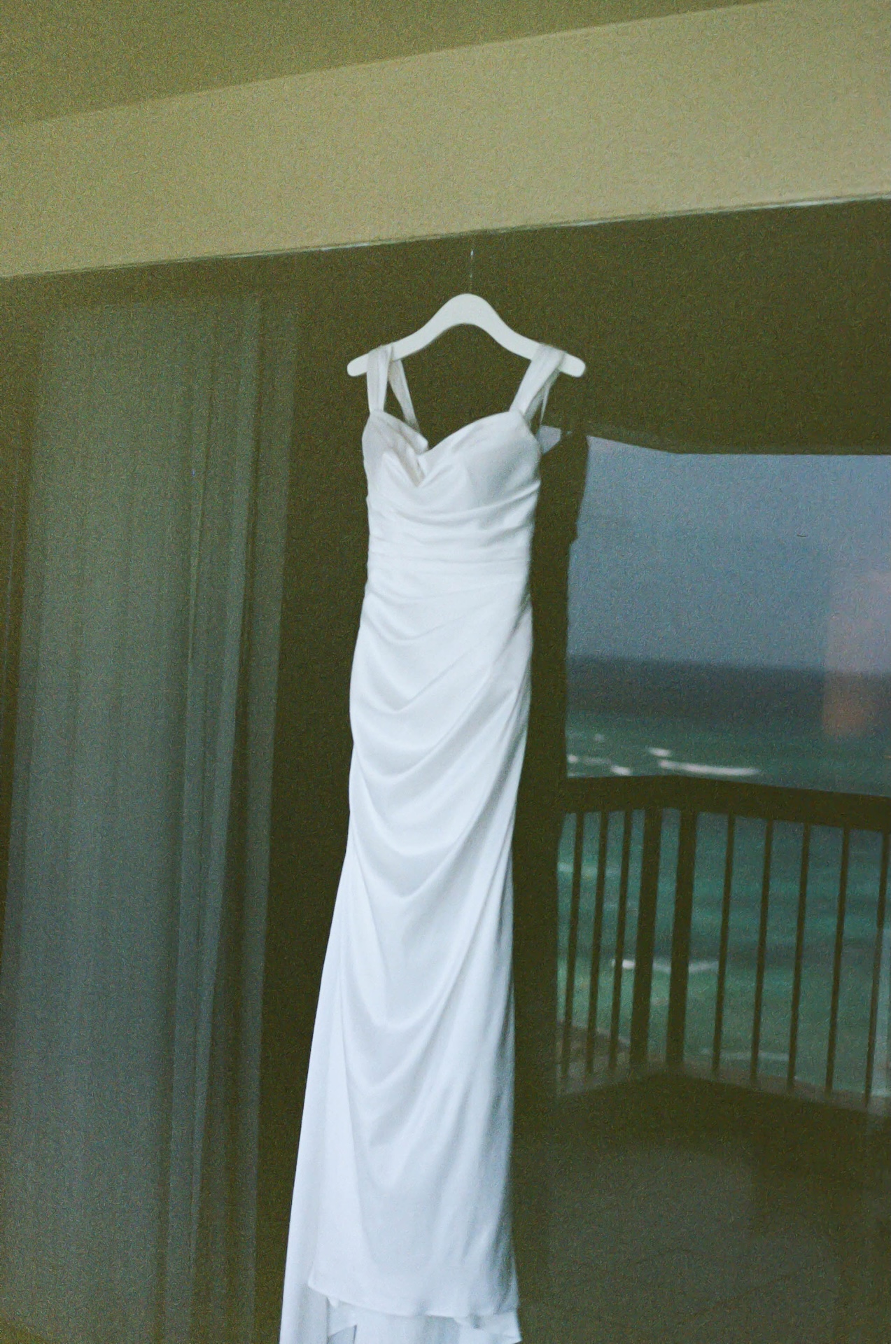 A wedding dress hangs in front of a balcony window, with soft ocean waves and sky reflected in the glass — a serene image captured by a destination wedding photographer.