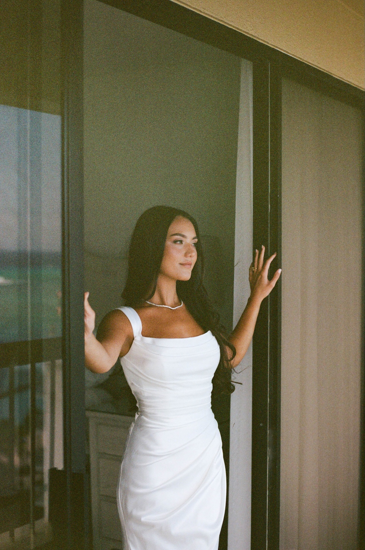 The bride stands confidently at a balcony doorway, holding open the glass panels as natural light highlights her gown and jewelry.