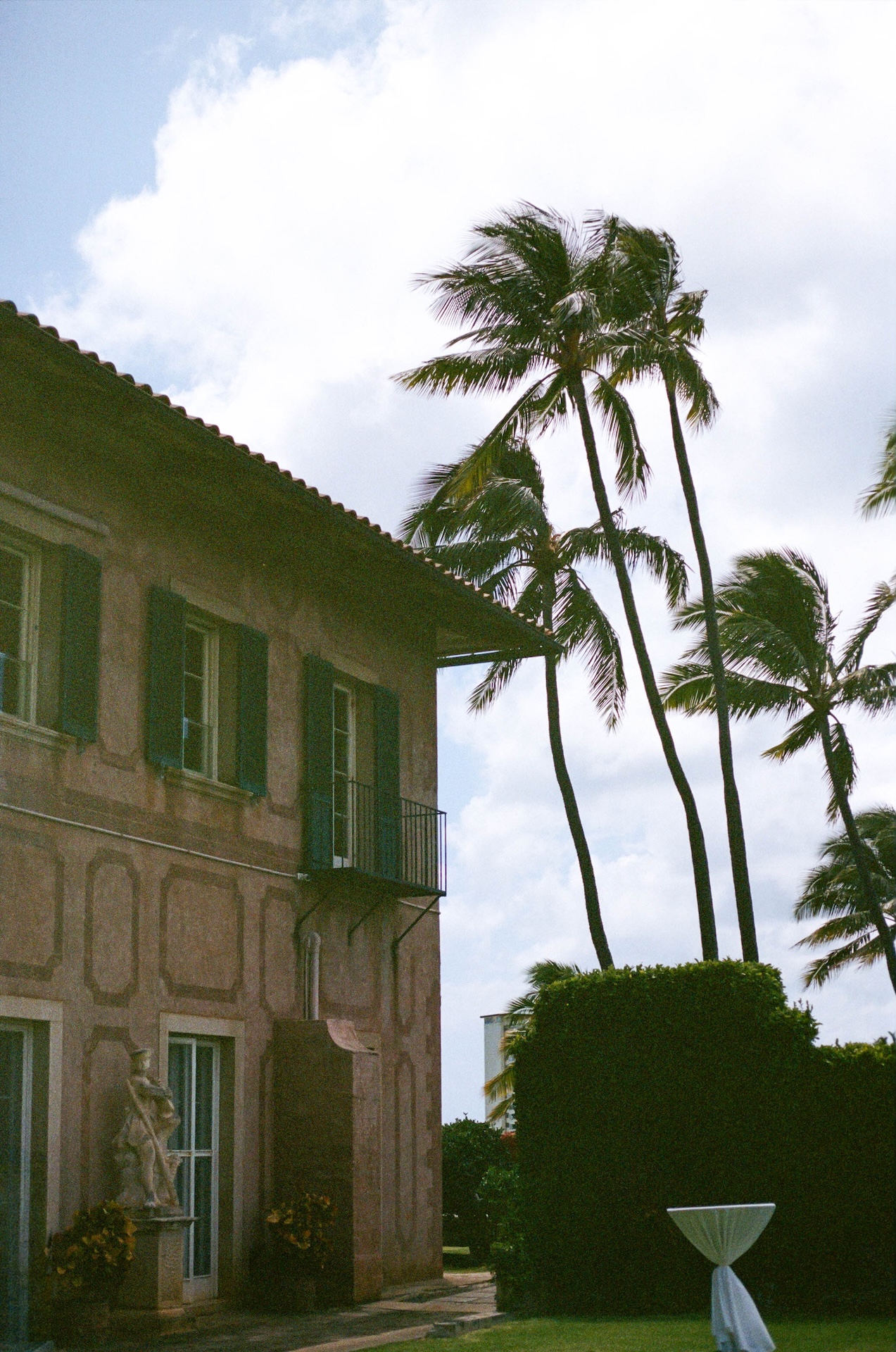 The side of a Mediterranean-style estate with a statue and tall palms rustling under a partly cloudy sky — a location scouted by a destination wedding photographer.