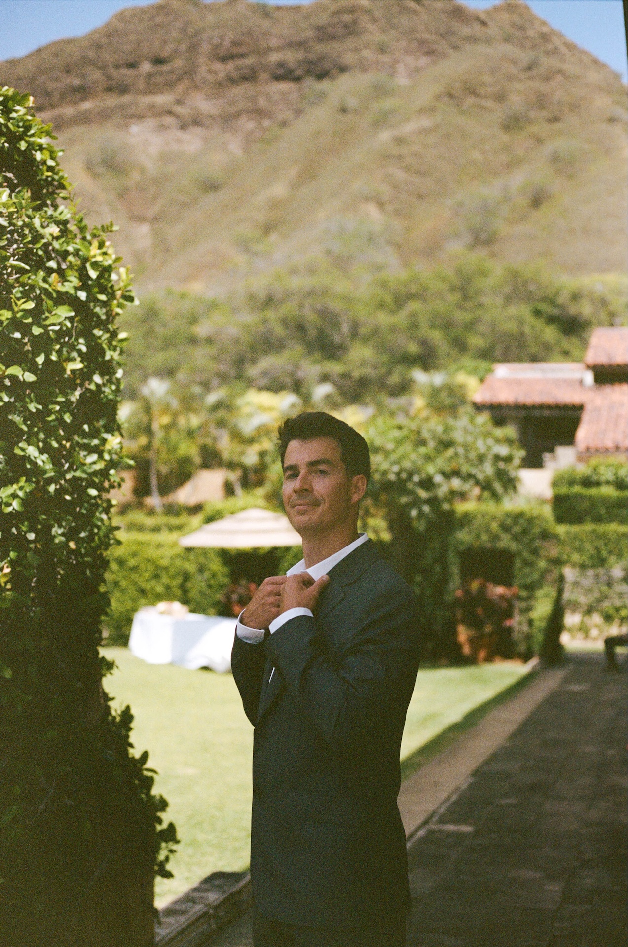 The groom straightens his collar while standing along a garden path, with mountains and manicured lawns behind him — a portrait by a destination wedding photographer.