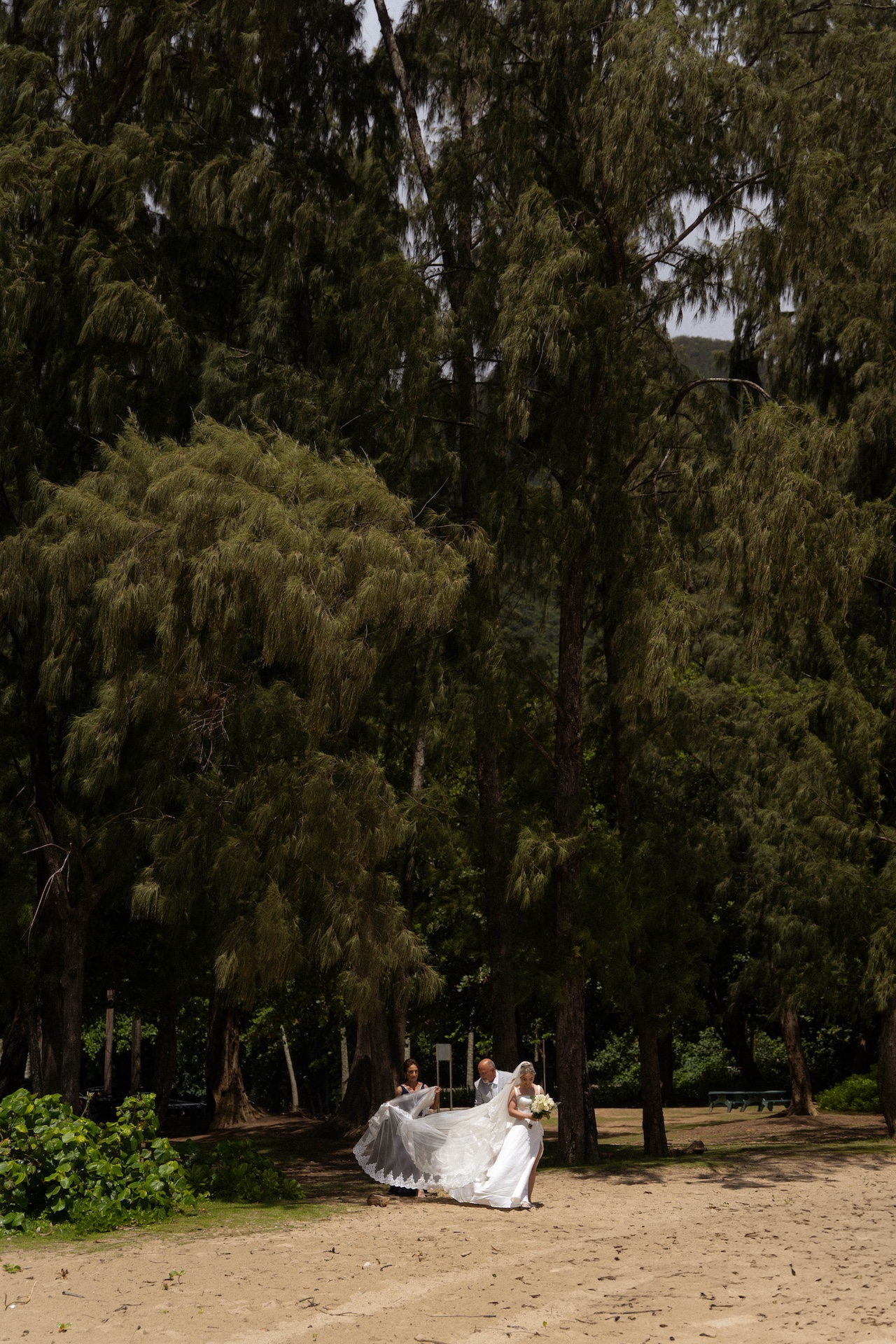 The bride's parents help her with her veil as she walks along the tree-lined beach path, holding her bouquet and trailing veil.
