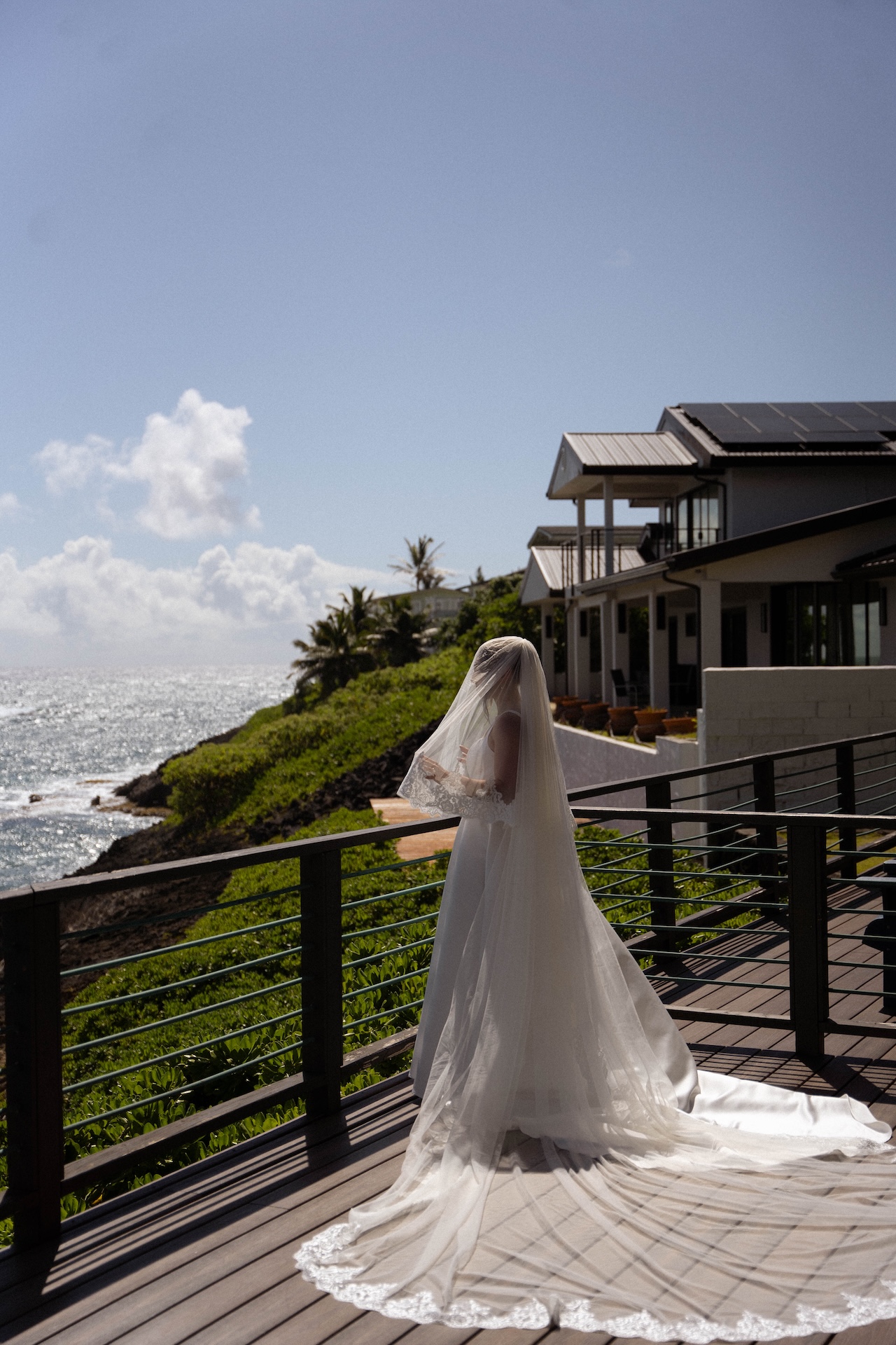 The bride, wearing a long lace veil and elegant gown, stands on a coastal balcony facing the ocean, quietly soaking in the peaceful pre-ceremony moment.