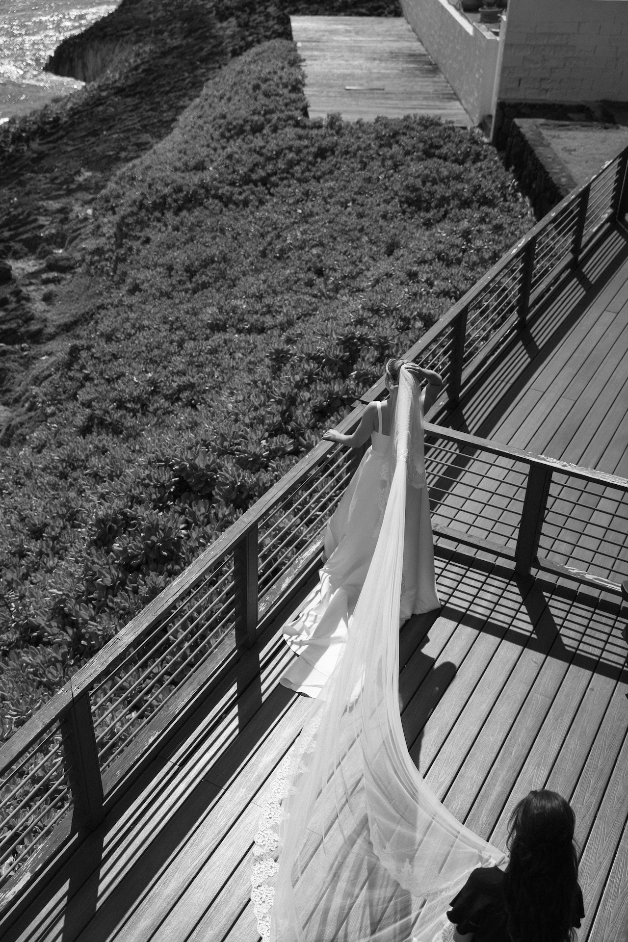 Overhead shot of the groom overlooking the Hawaii waves and ocean while she holds her veil and her mom fixes it for her. 