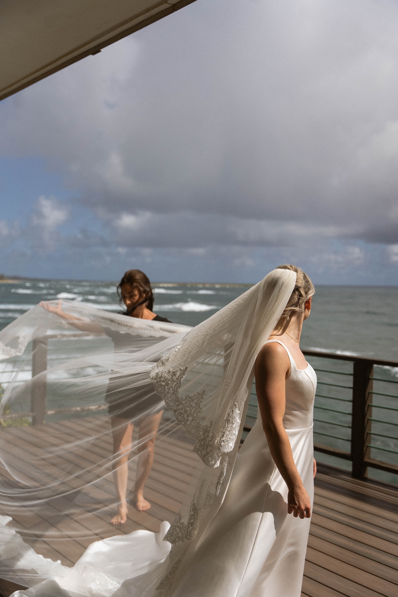 The bride’s veil flows in the wind as her mom helps arrange it on a breezy balcony with ocean views.