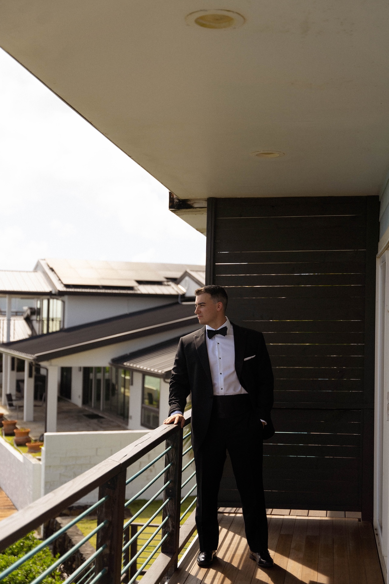 Groom stands alone on a balcony in a black tuxedo, looking out thoughtfully before the ceremony at their scenic airbnb wedding location in Hawaii.