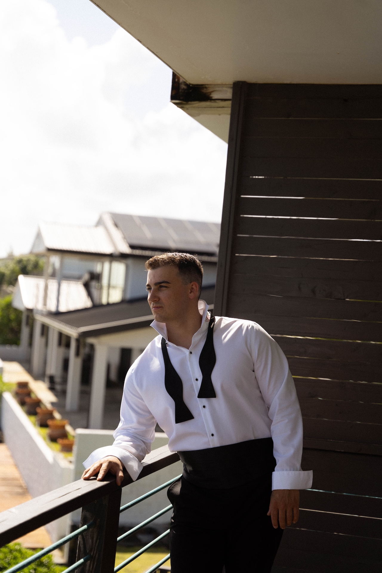 The groom leans against the railing at the airbnb as he overlooks the ocean while getting ready.