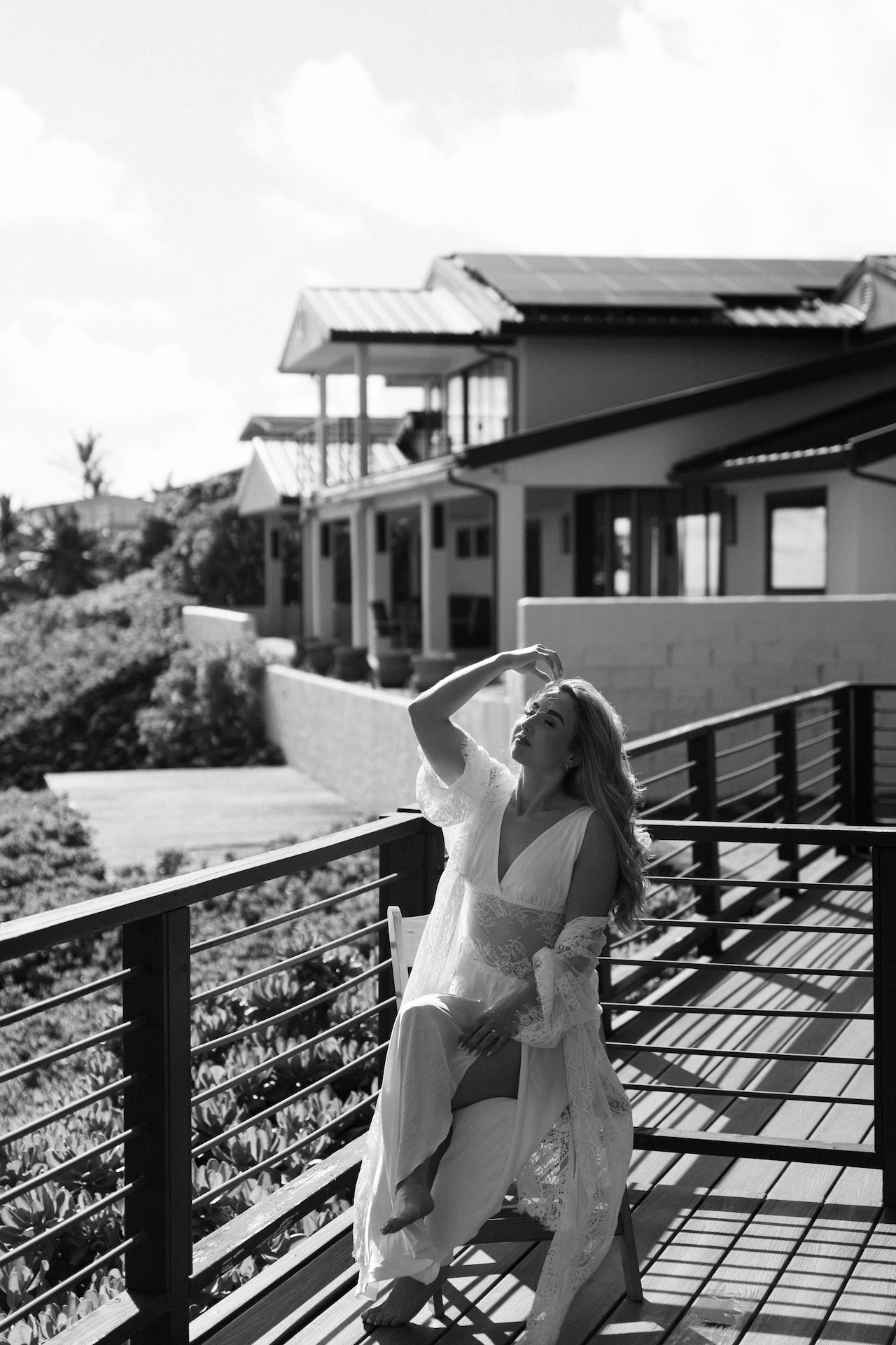 Bride in a lace robe sits on the balcony, smiling and brushing her hair back as sunlight pours over the airbnb wedding venue.