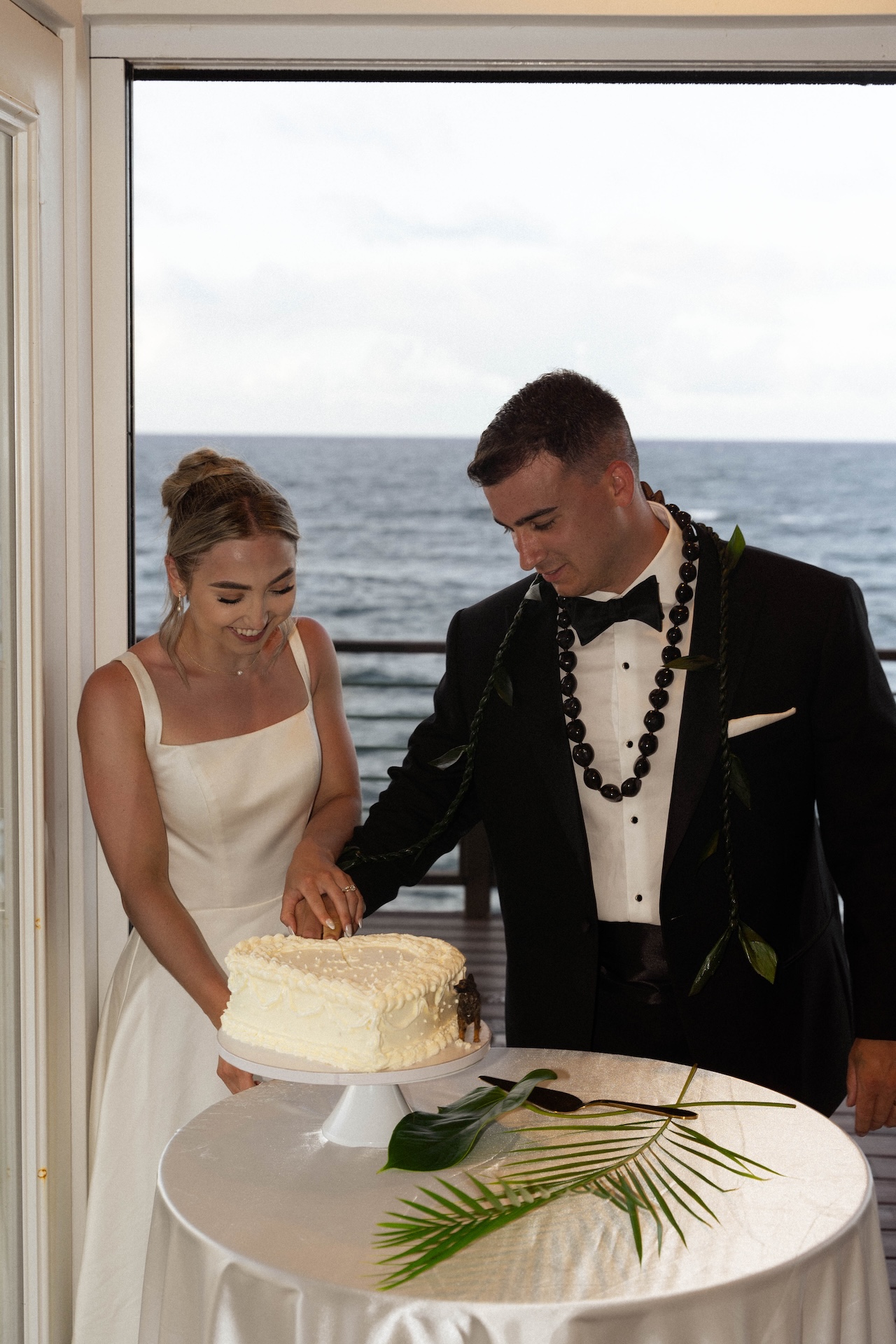 The couple smiles while cutting their cake indoors, celebrating their intimate airbnb wedding with views of the sea behind them.