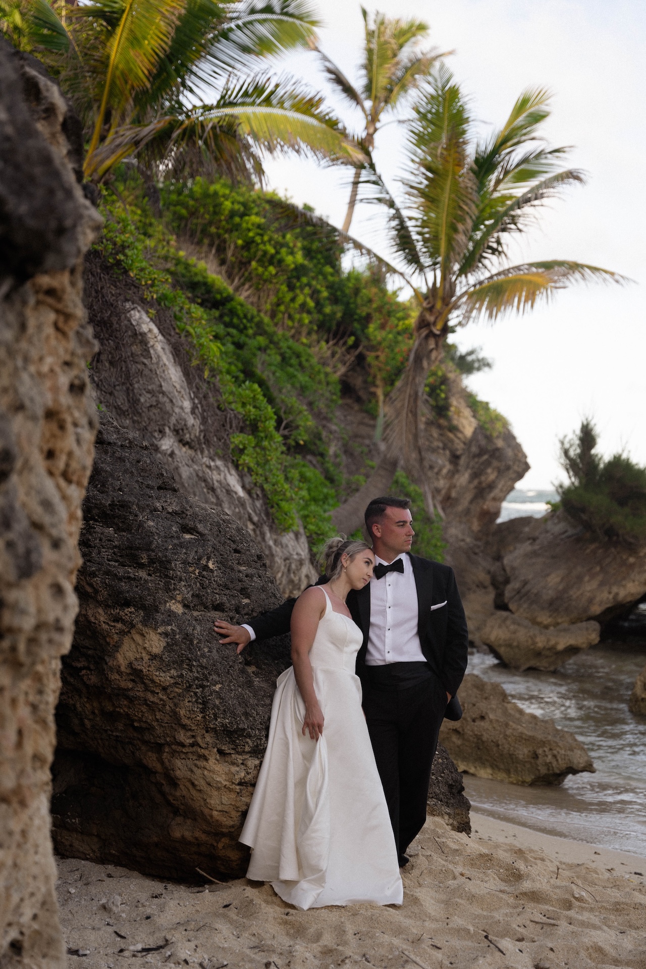 Bride and groom pose against seaside cliffs, framed by tropical palm trees during their romantic airbnb wedding portrait session.