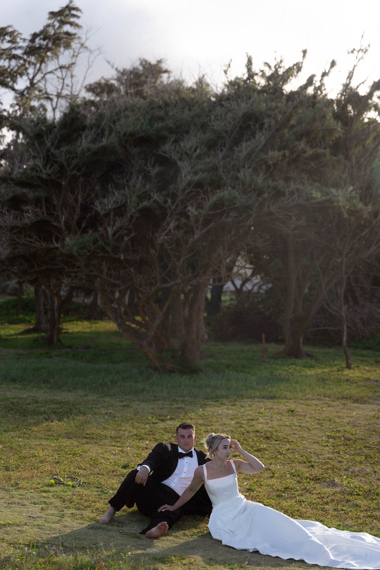 The bride and groom sitting on the grass at their airbnb wedding with big trees behind them as they stare out at the beach.