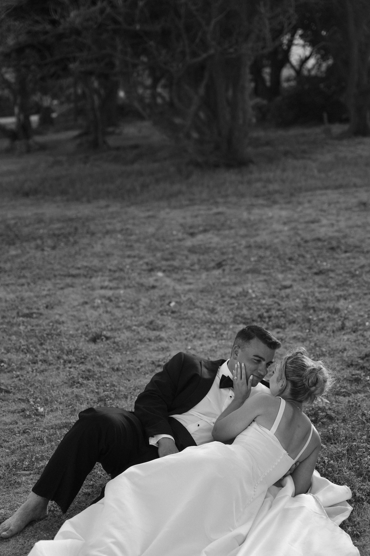 Bride and groom laying on the grass while the bride cradles the grooms face as they stare at each other lovingly.