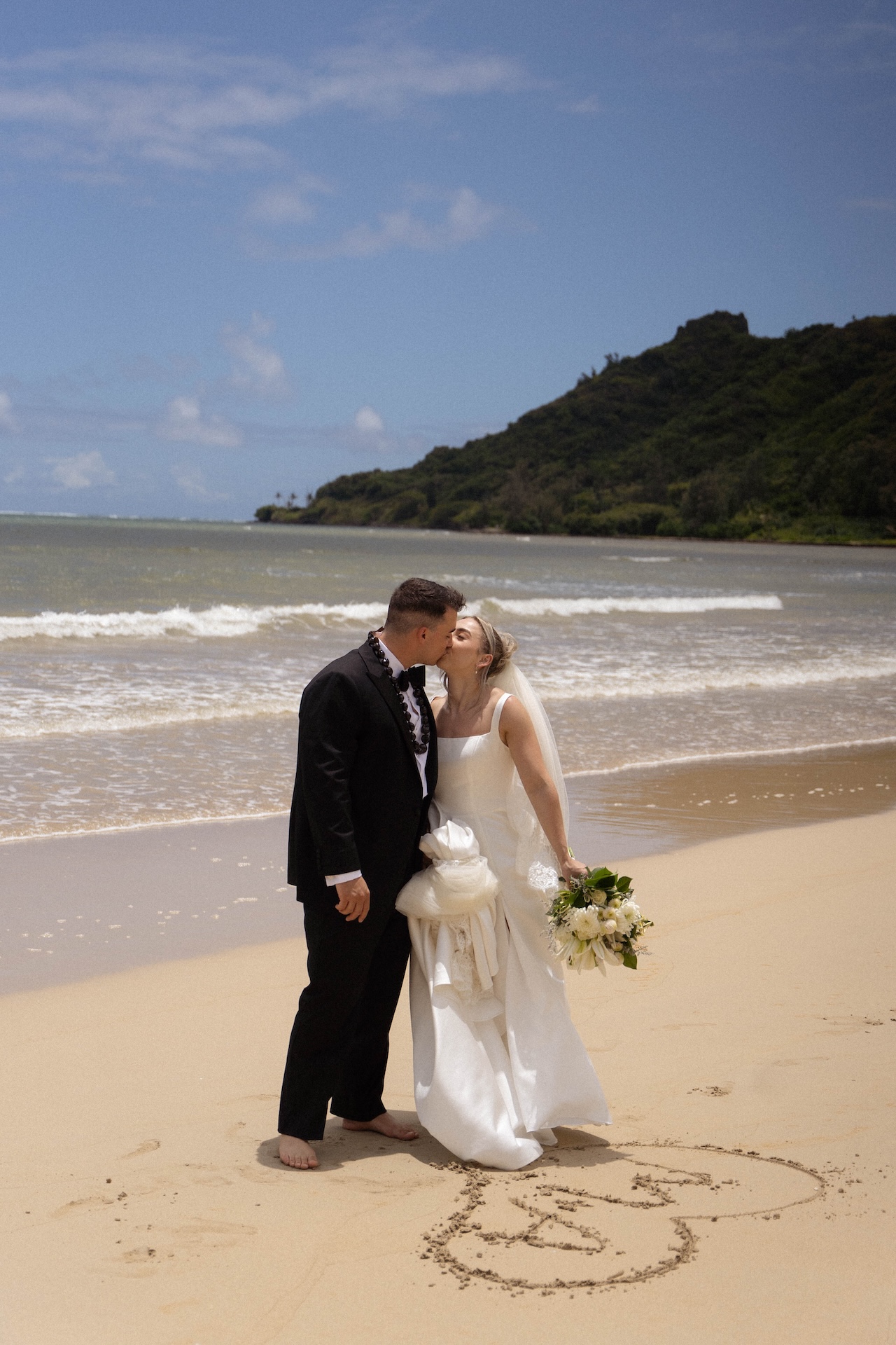The bride and groom share a kiss on the beach while standing behind their intials and a heart they drew in the sand. 