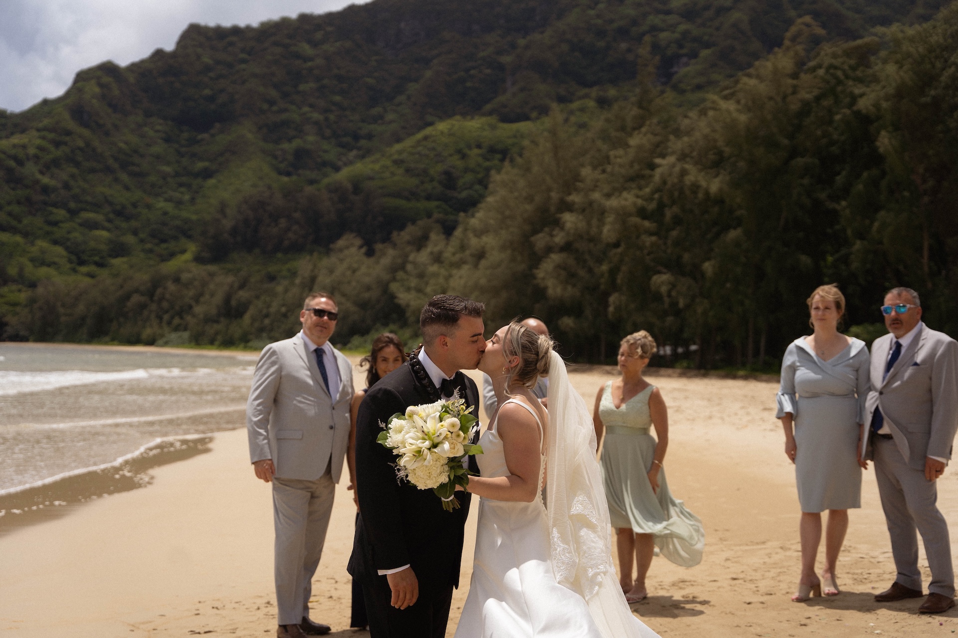 The bride and groom share a kiss on the beach surrounded by family, capturing the heartfelt moment of their airbnb wedding ceremony.