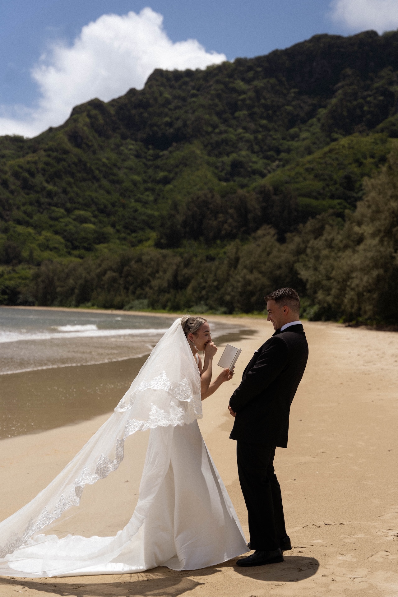 The bride reads her vows with joyful emotion while standing on a sandy beach facing her groom during their intimate airbnb wedding on the Hawaiian coast.