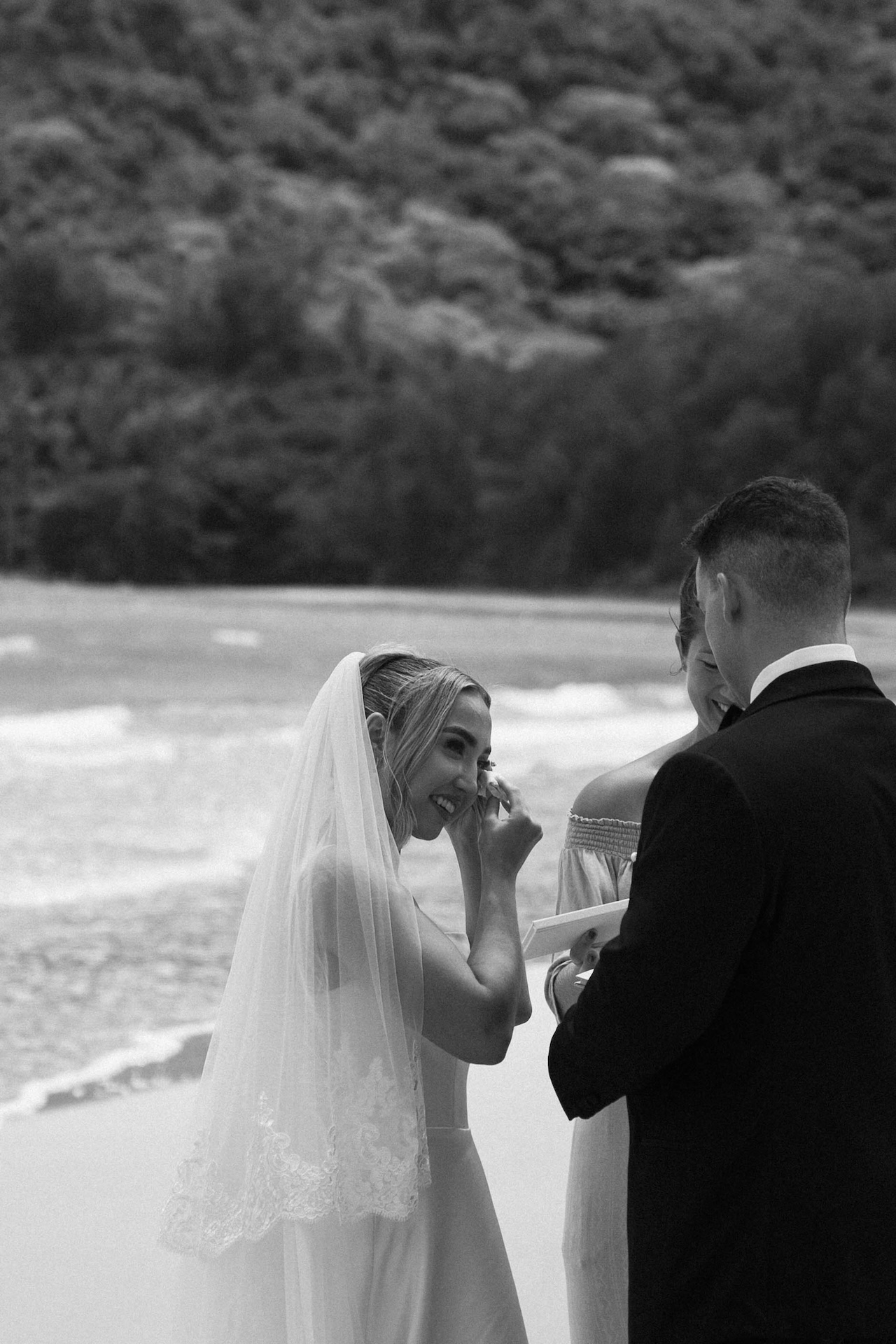 Tearful and smiling, the bride wipes her face during the vow exchange, surrounded by ocean waves and soft wind in a deeply emotional ceremony moment.