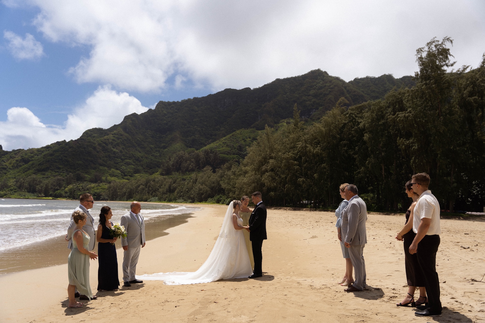 Small wedding ceremony on a secluded beach with loved ones standing on either side as the couple exchanges vows beneath a backdrop of lush mountains.