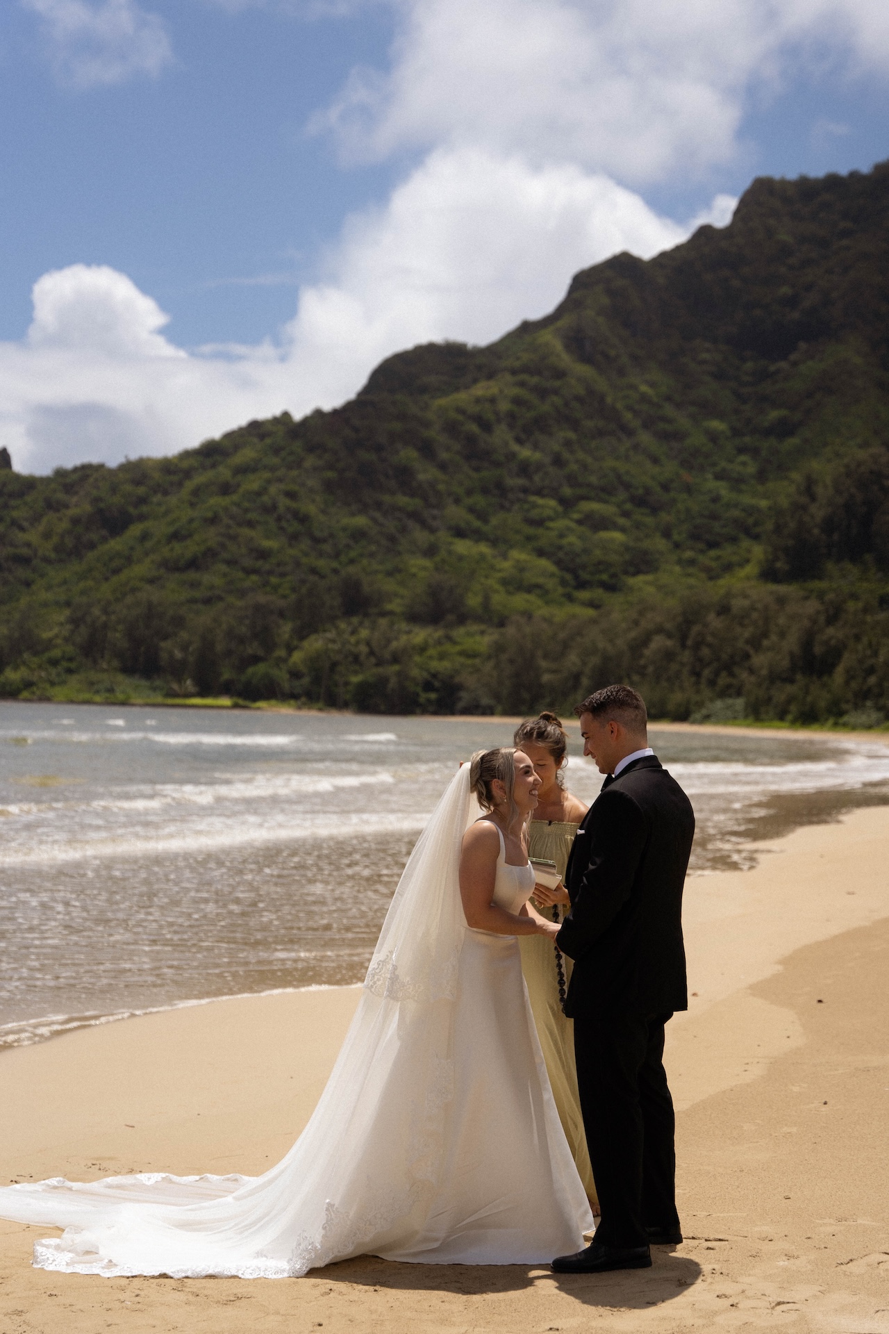 The bride and groom laughing and holding hands on the beach as the officiant reads their vows to them. 
