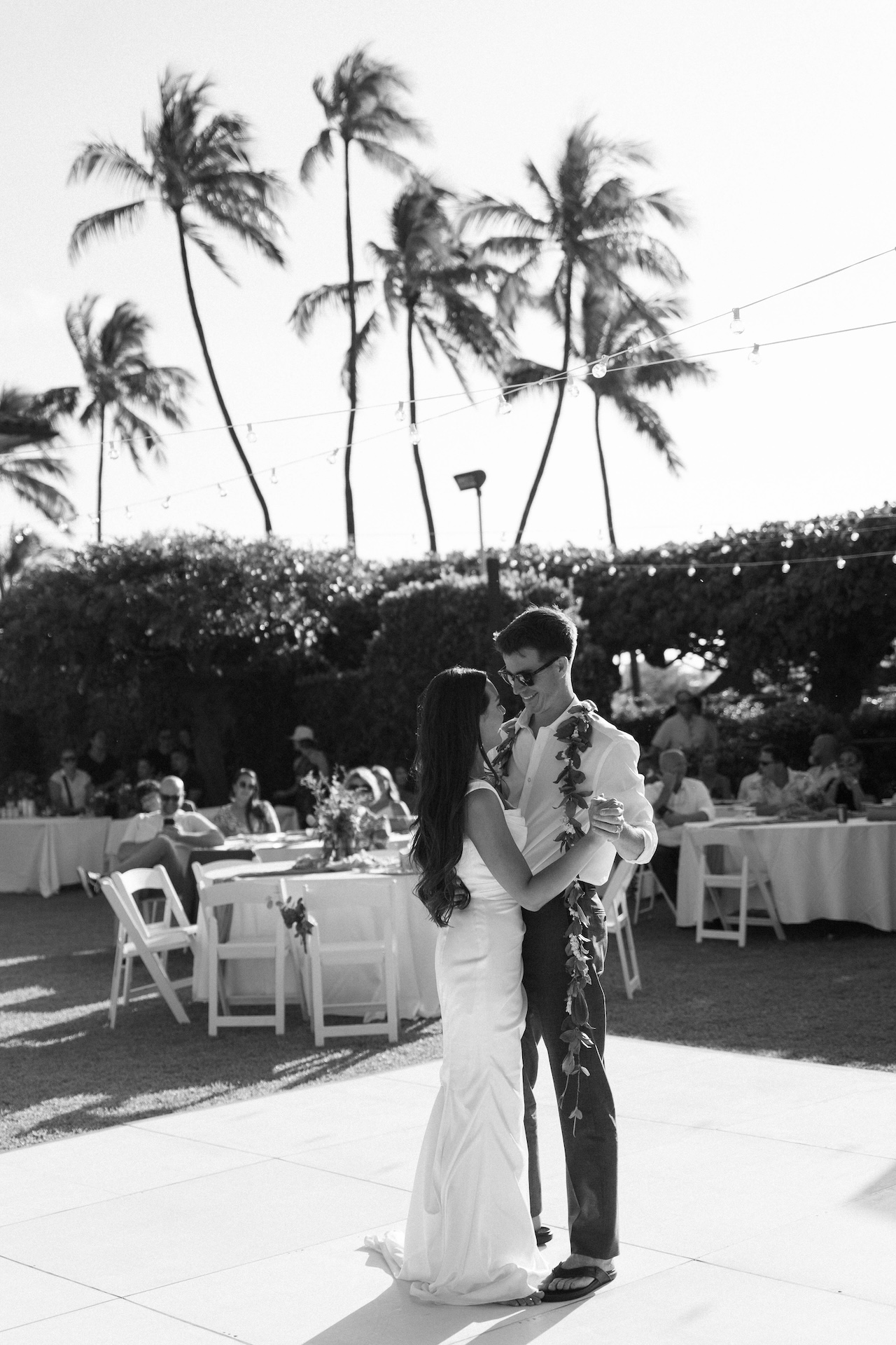 Black and white photo of the bride and groom smiling during their first dance, with guests seated beneath palm trees.