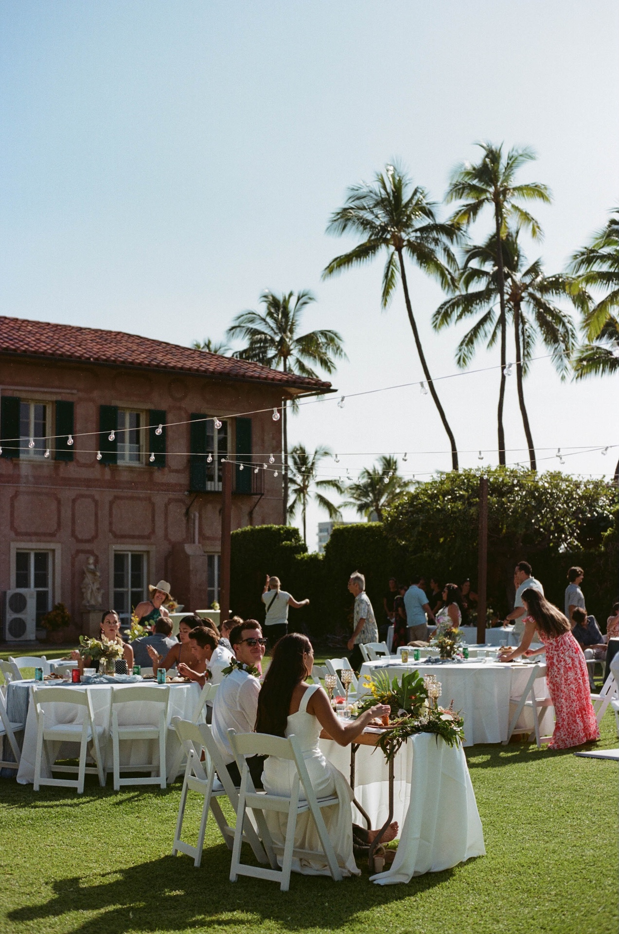 Guests mingle and dine at white tables set on a sunny lawn with palm trees swaying behind a Tuscan-inspired villa — captured by a destination wedding photographer.