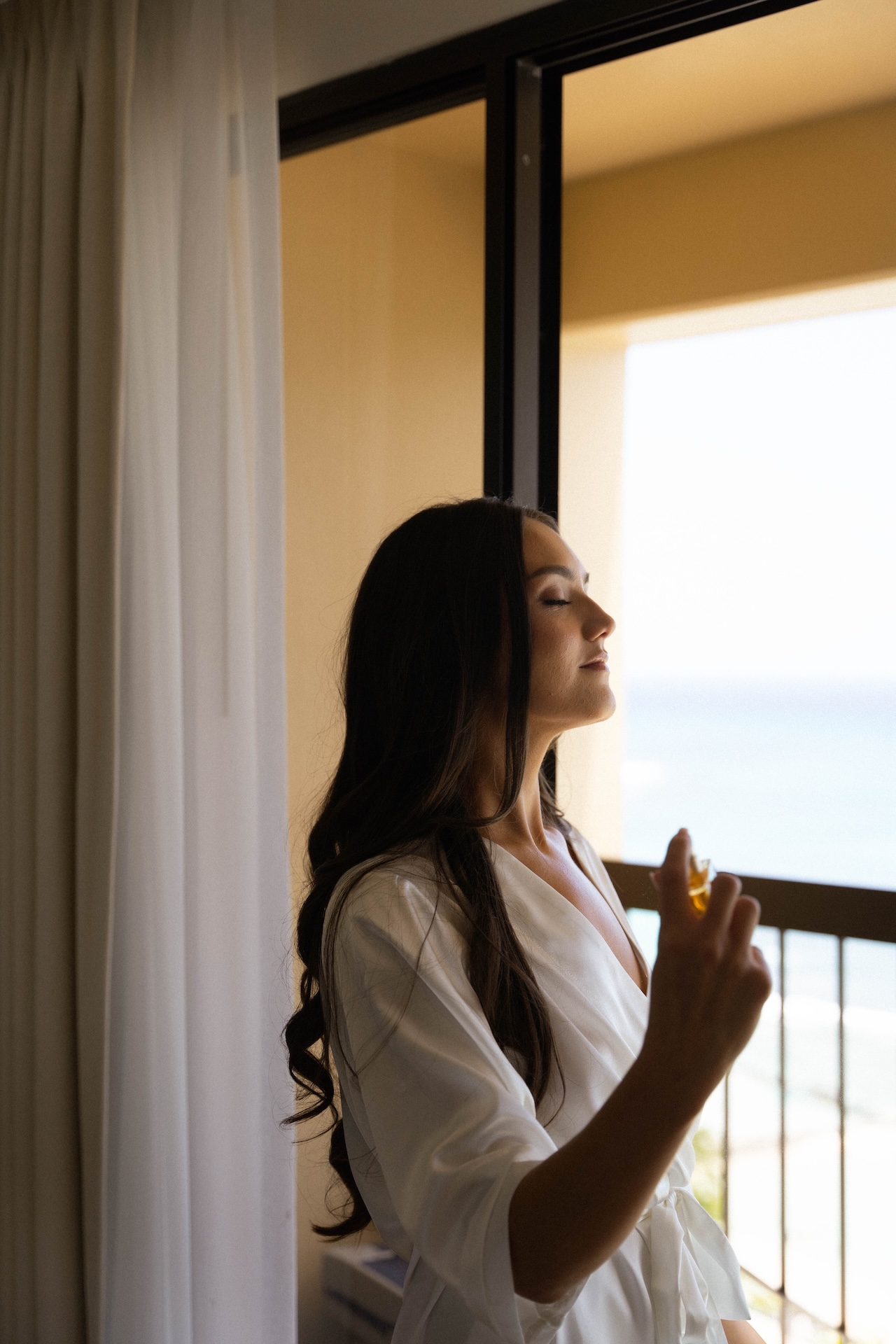 The bride closes her eyes as she sprays perfume on her neck, soft morning light falling through a nearby curtain — captured by a destination wedding photographer.