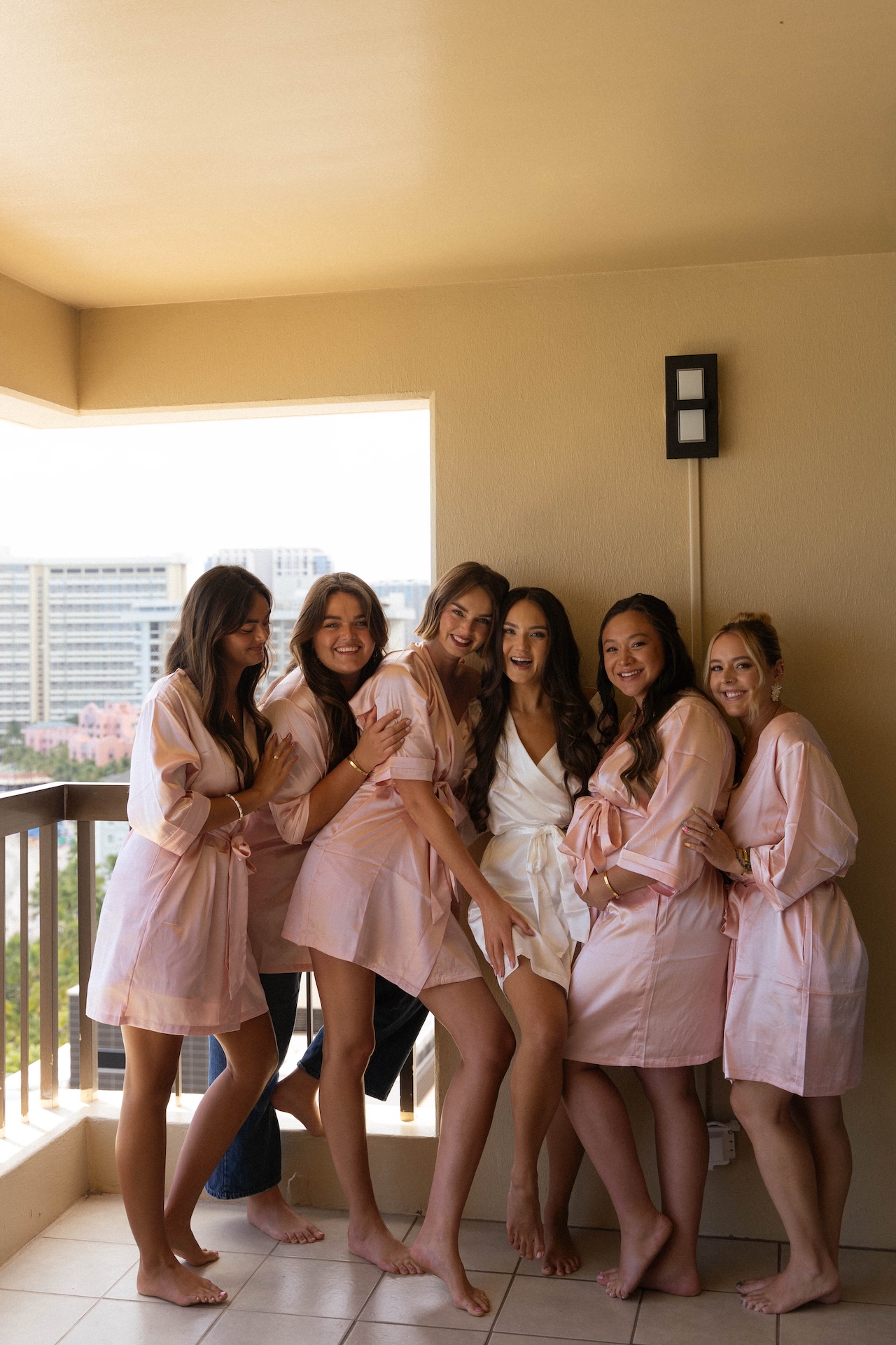 The bride poses on a balcony with her bridesmaids in blush pink robes, all laughing and barefoot on tiled flooring.