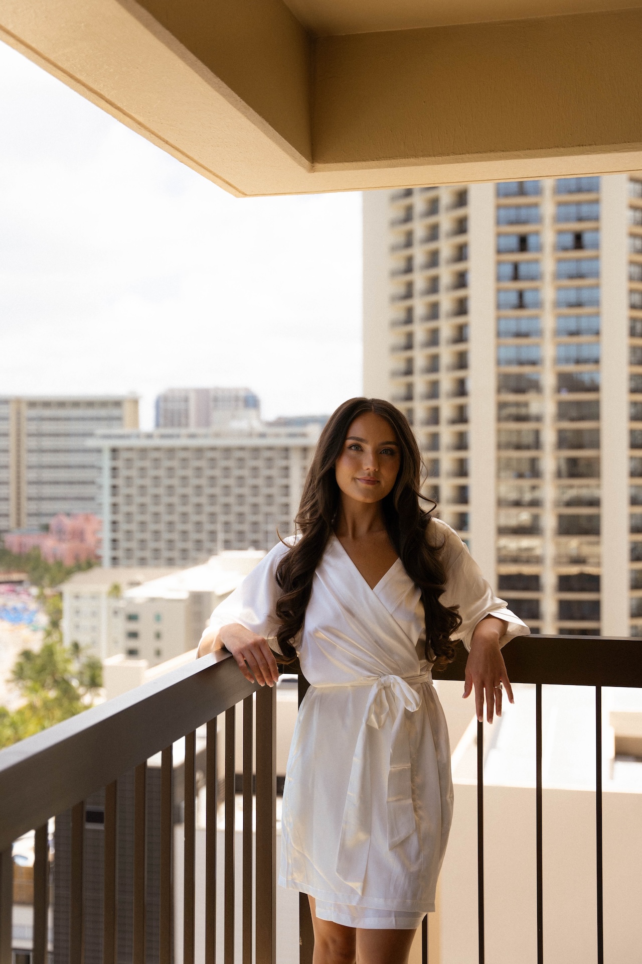 The bride poses on a high-rise balcony in a white robe, city buildings and Waikiki in the distance.