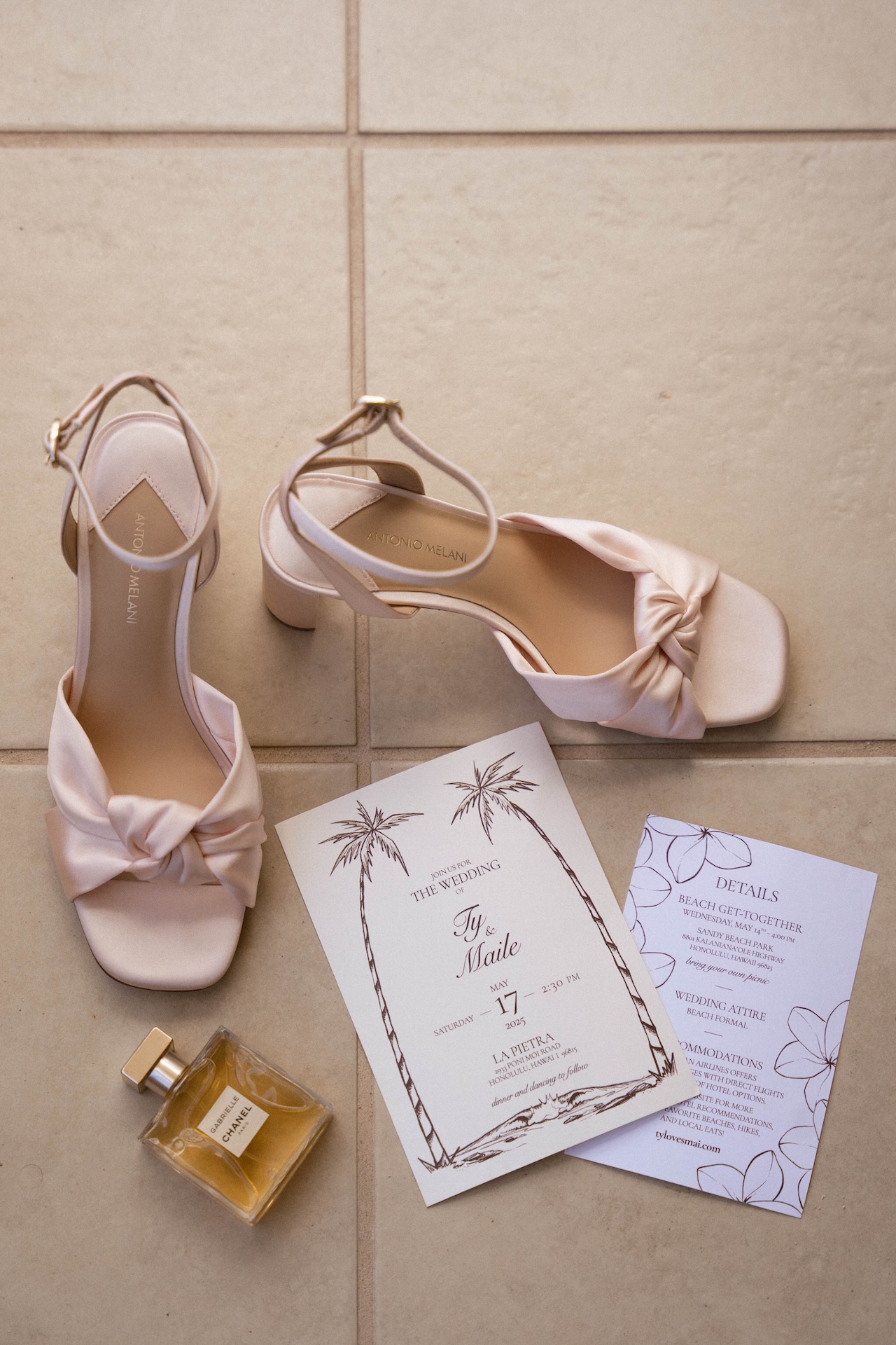Flat lay of wedding details including blush pink heels, perfume, and the couple's palm tree-themed invitation — styled by a destination wedding photographer.