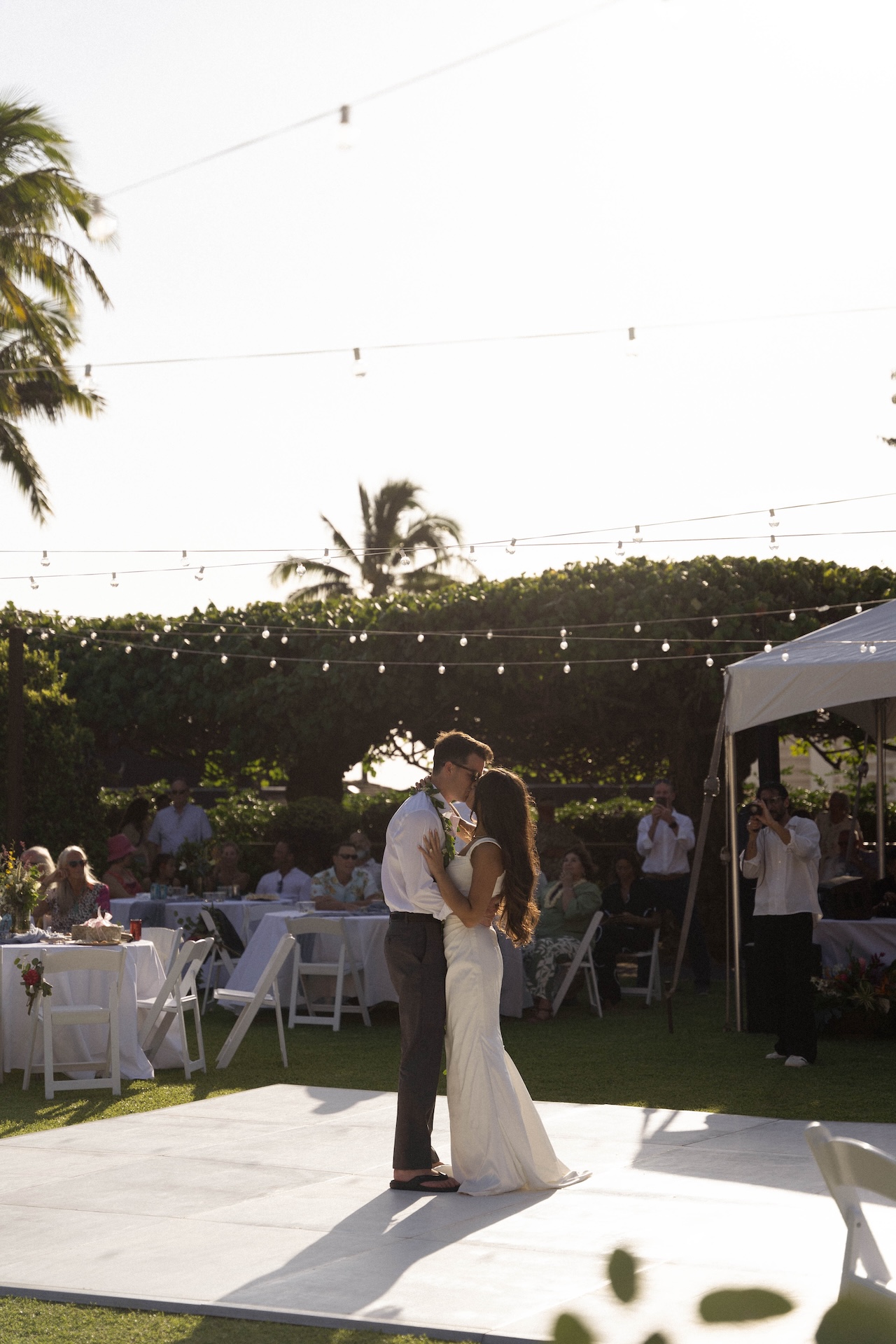 The bride and groom share a soft kiss on the dance floor during their first dance, sunlight casting long shadows across the ground.