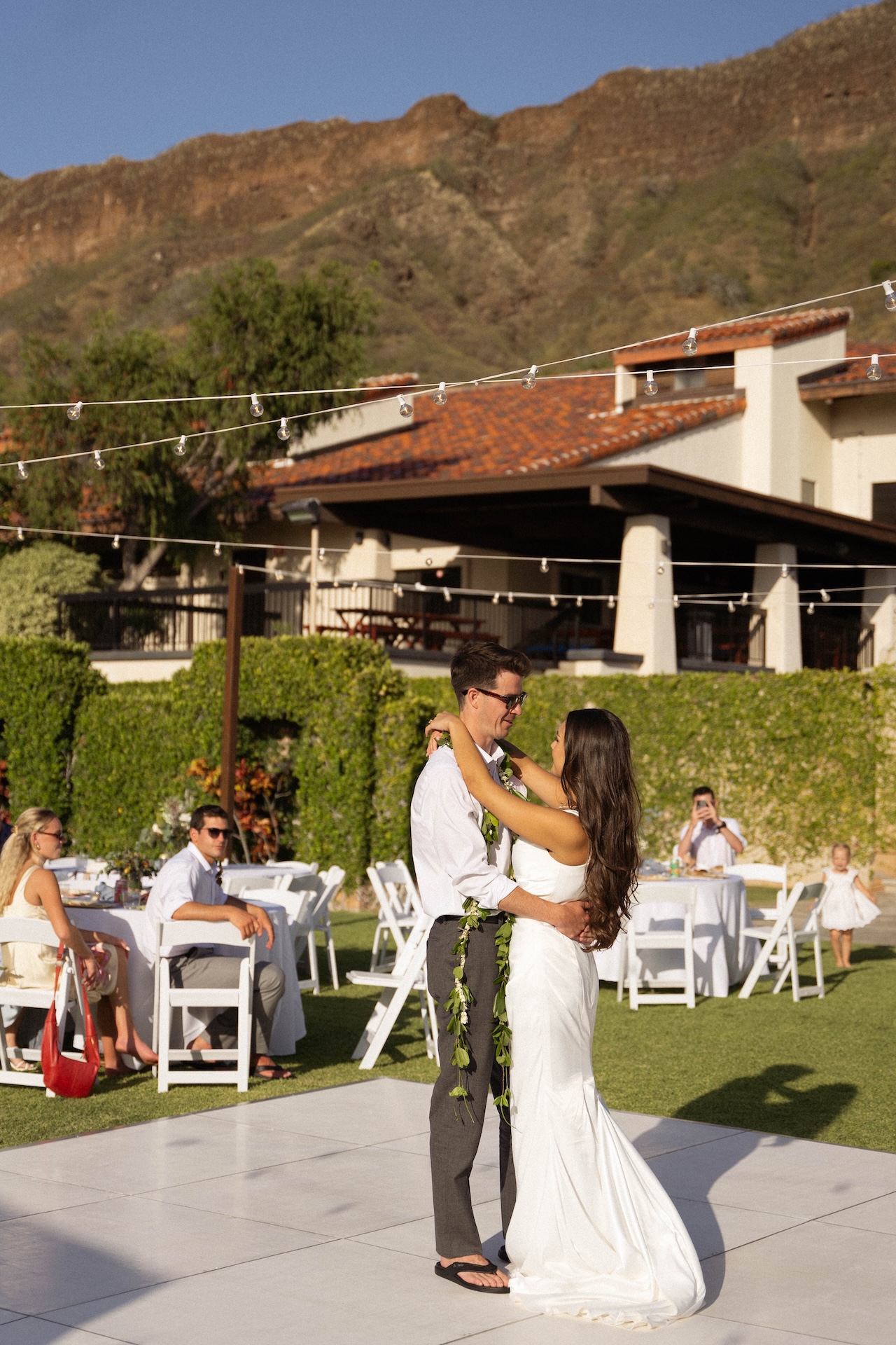 The couple slow dances at golden hour on a white dance floor, the mountains rising behind them in the distance.
