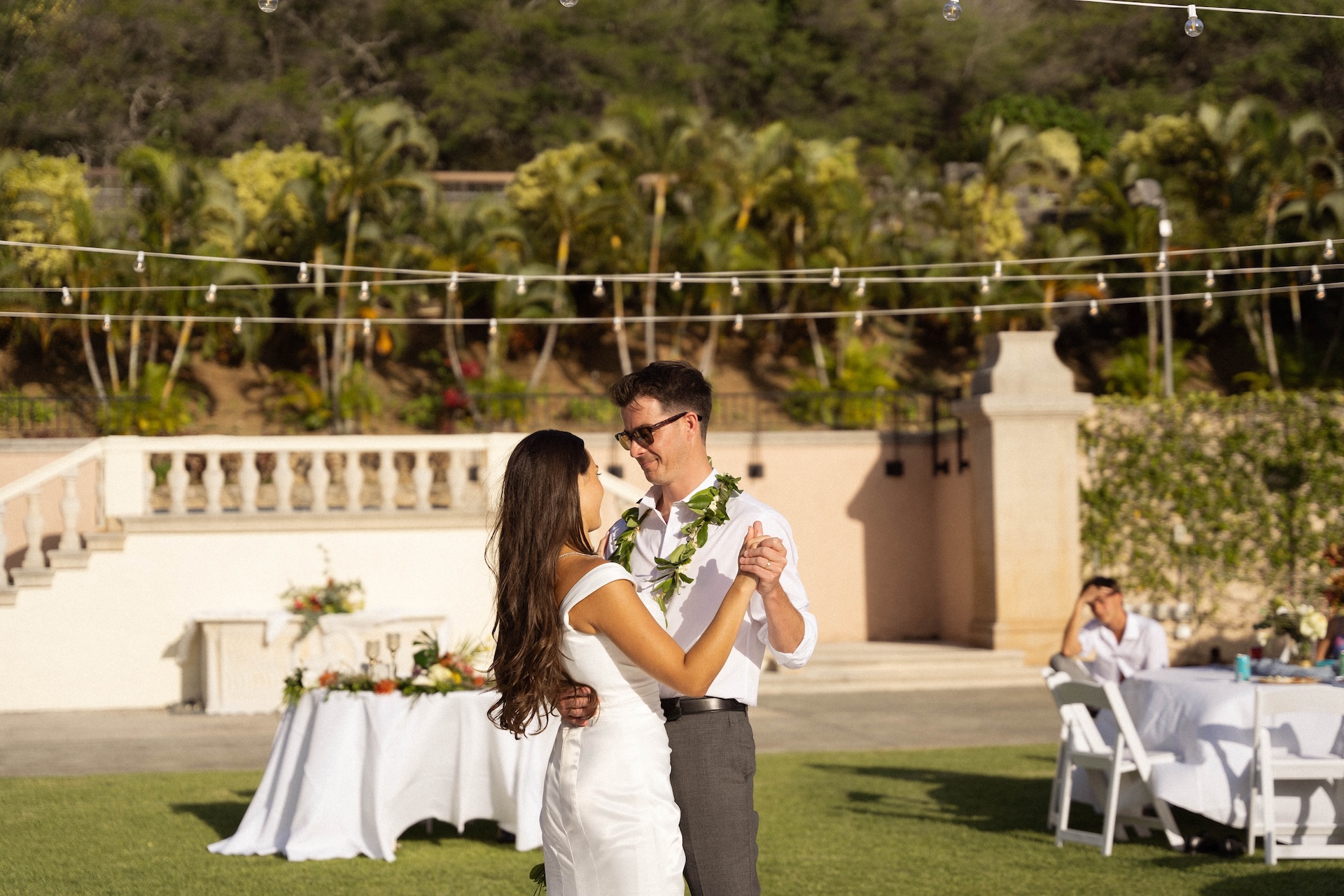 The couple dances under café lights on a grass lawn with a Mediterranean-style estate in the background — a romantic scene by a destination wedding photographer.