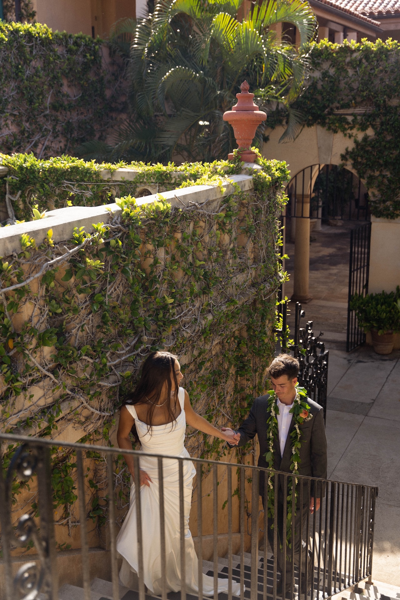 The groom holds the bride’s hand gently as he leads her up vine-covered courtyard stairs — a golden hour scene by a destination wedding photographer.