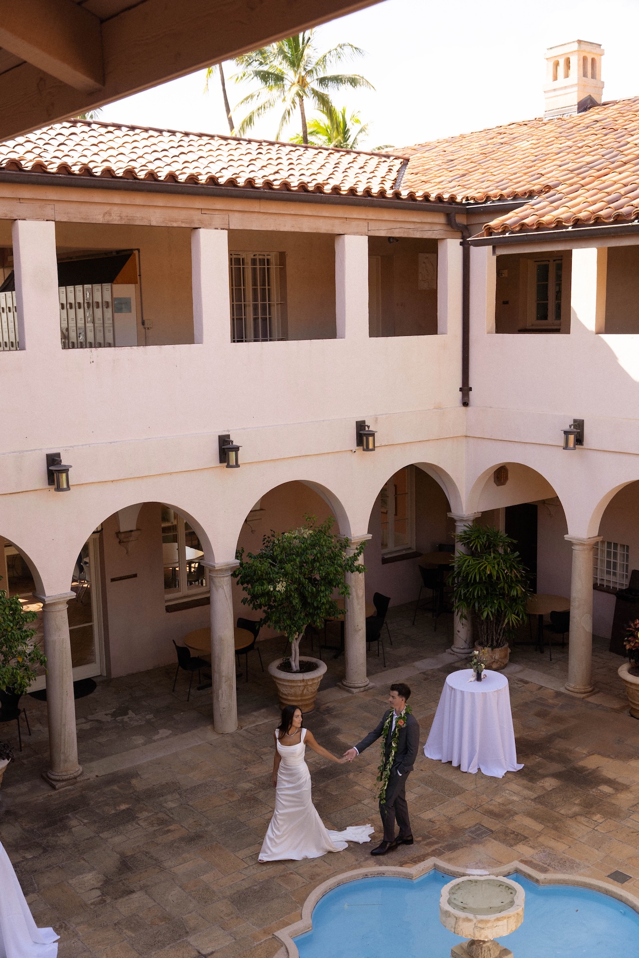 A wide shot of the couple in an arched courtyard by a fountain, holding hands as they take in the view.