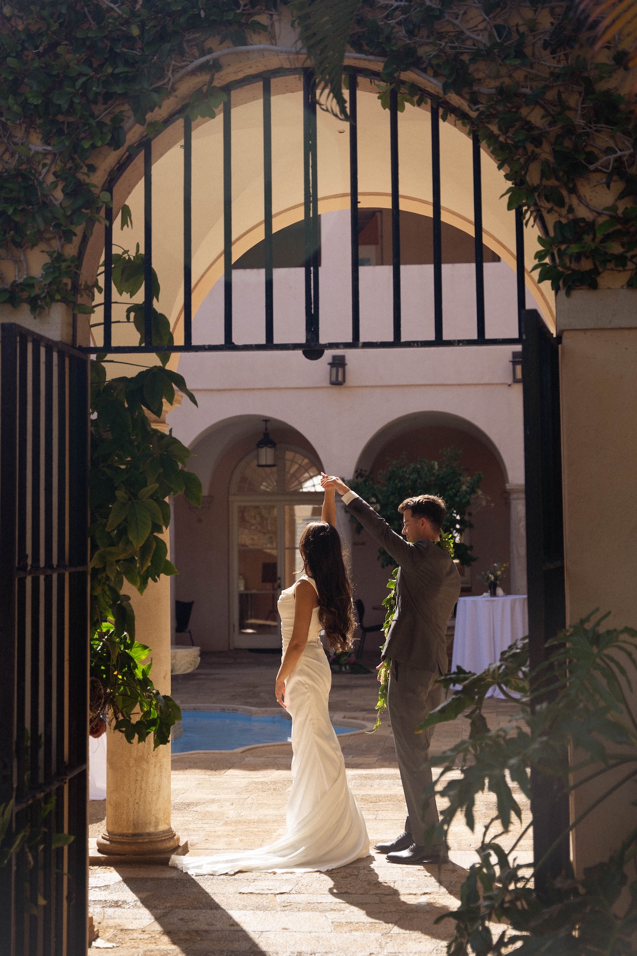 The couple dances under a gated archway, sunlight streaming through as they twirl in a romantic courtyard.