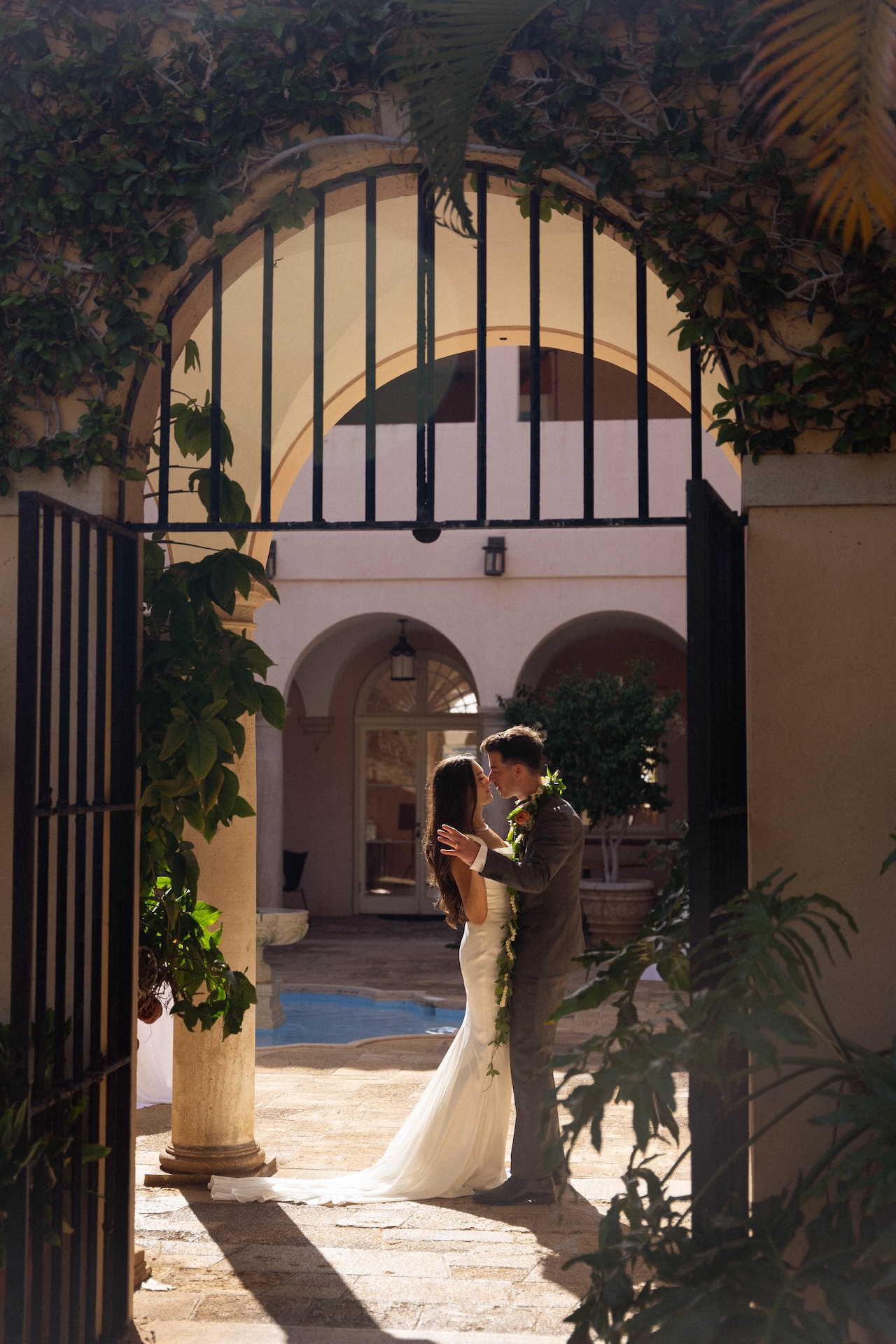 The couple embraces beneath a grand arched gate draped in greenery, bathed in golden light — photographed by a destination wedding photographer.