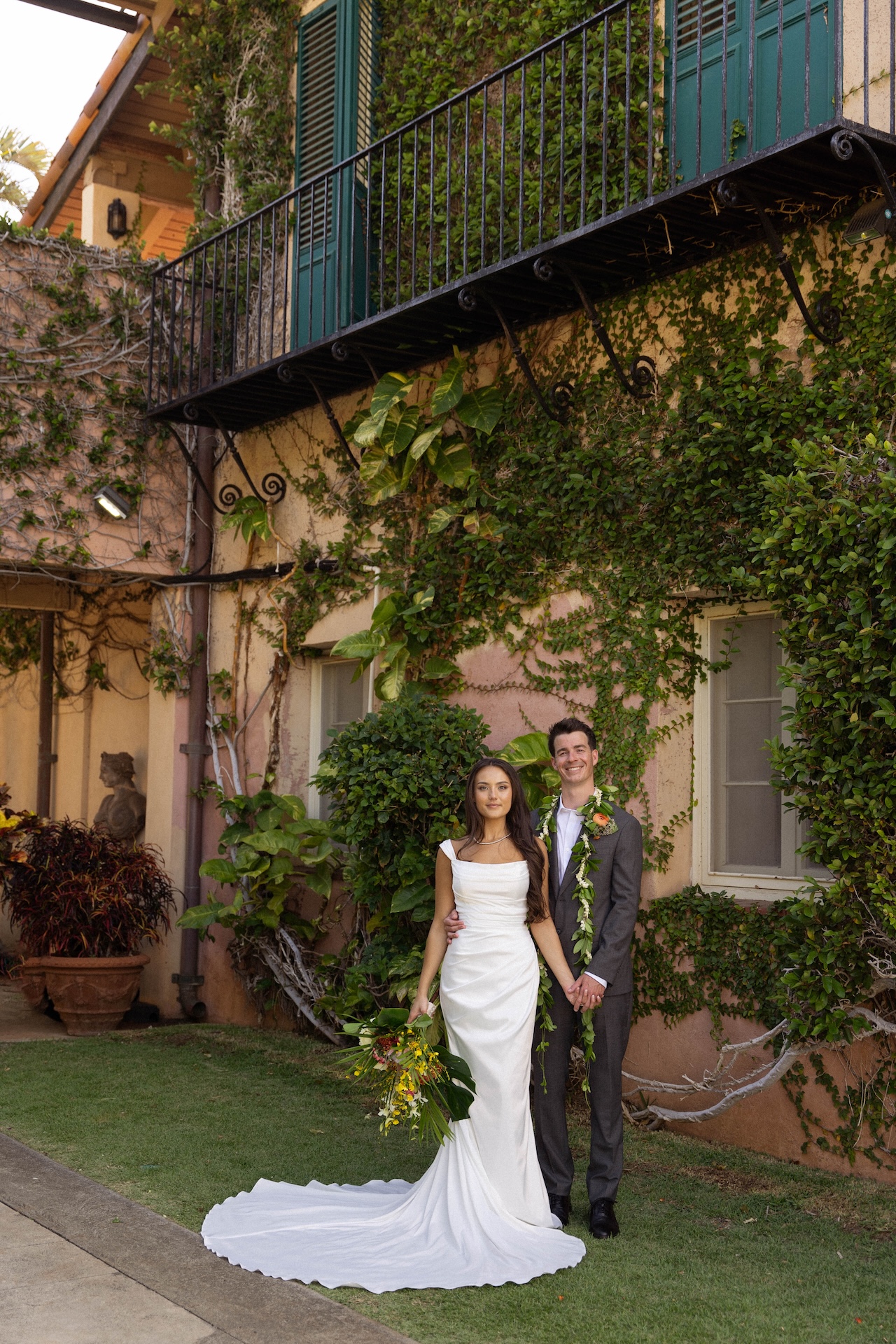 A bride and groom stand hand-in-hand in front of a vine-covered villa wall, with the bride’s train elegantly flowing on the grass.