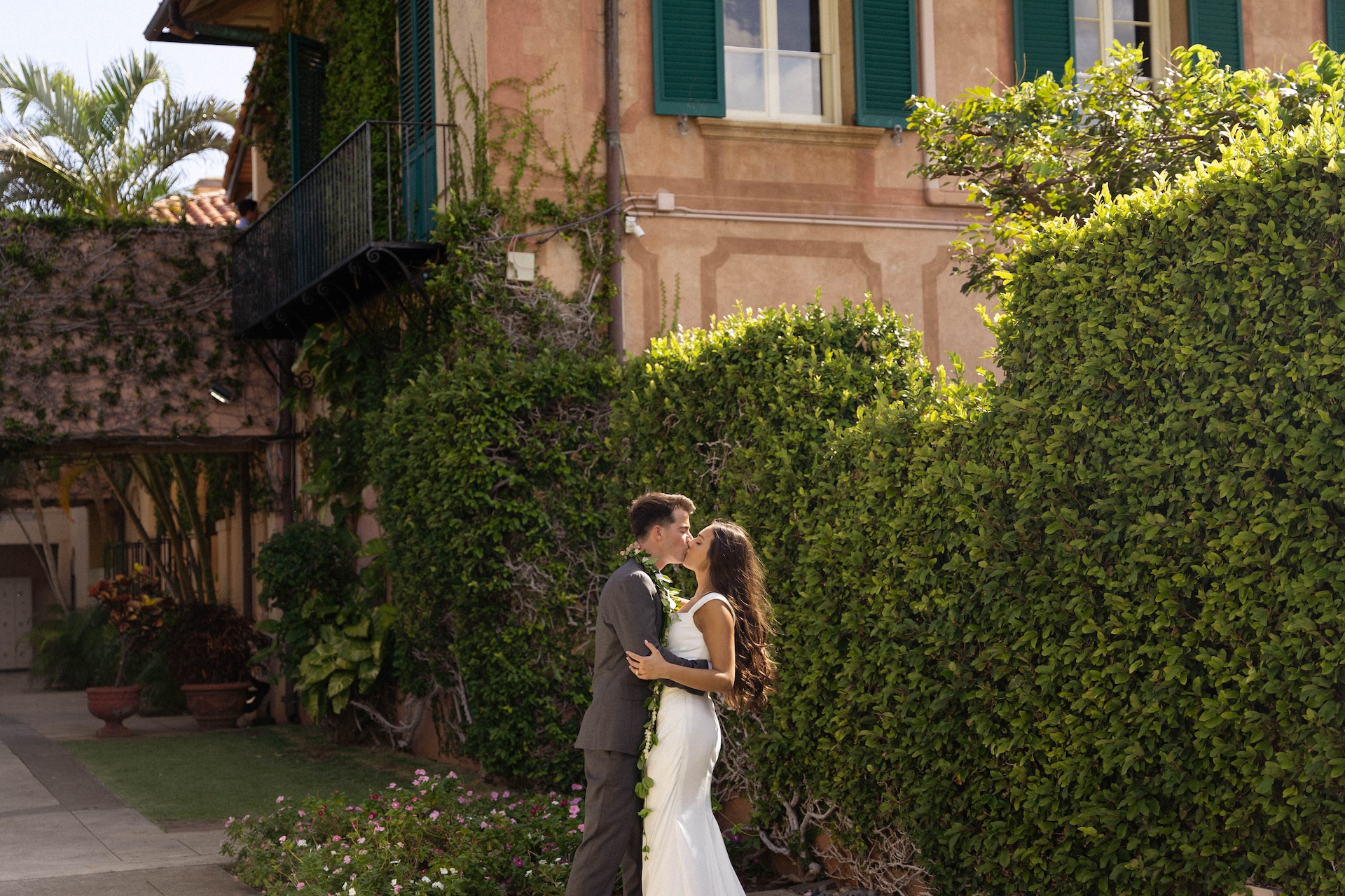 A tender kiss between the couple as they stand by manicured hedges and a Mediterranean-inspired building, captured by a destination wedding photographer.