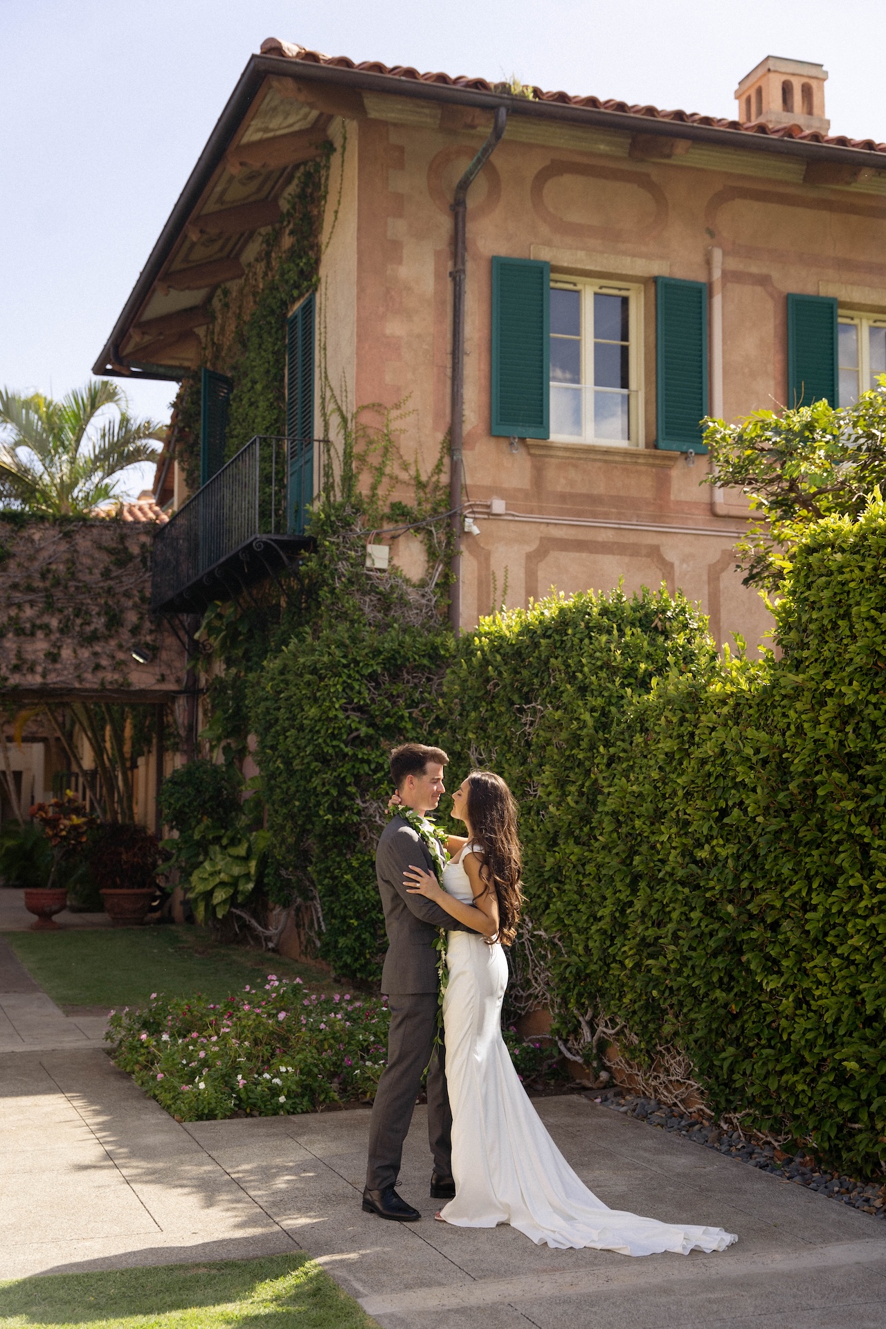 The couple stands forehead-to-forehead, embraced in front of a villa covered in ivy and green shutters — captured by a destination wedding photographer.