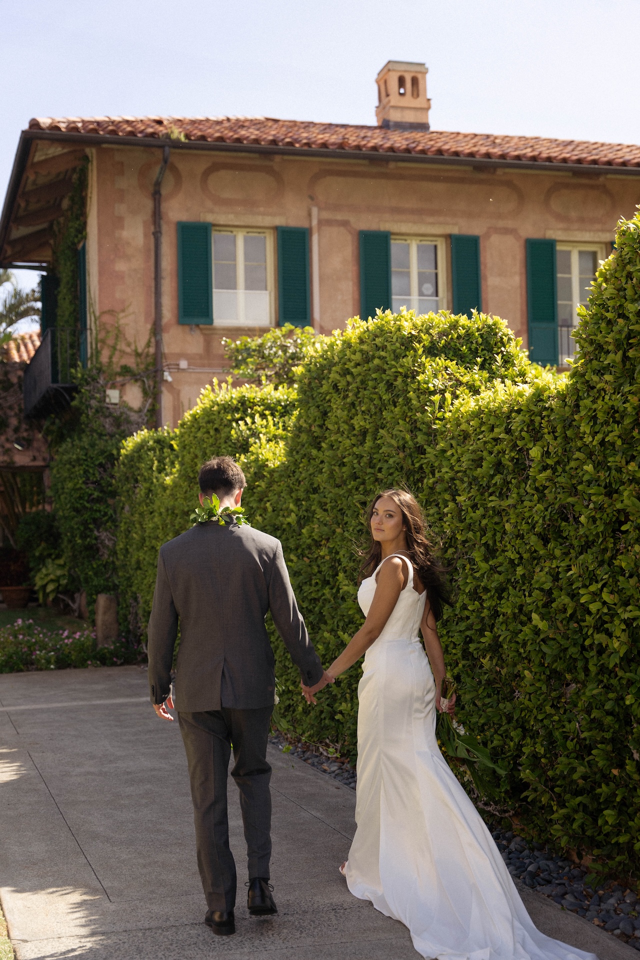 The bride looks back while walking hand-in-hand with the groom along a garden pathway framed by green hedges and a Mediterranean-style villa.