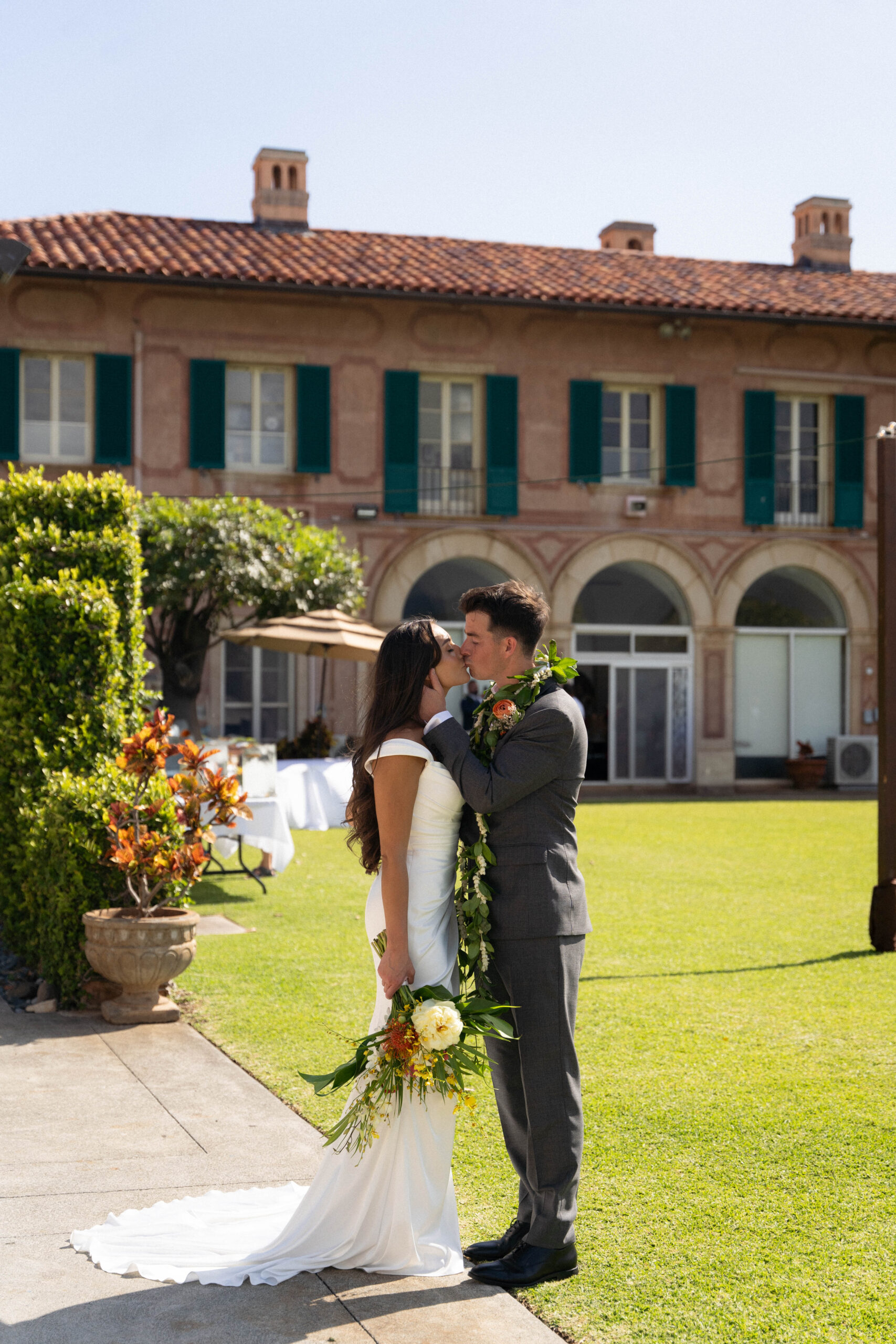 A romantic kiss between the couple in front of an old-world villa, the bride holding a tropical bouquet — captured by a destination wedding photographer.
