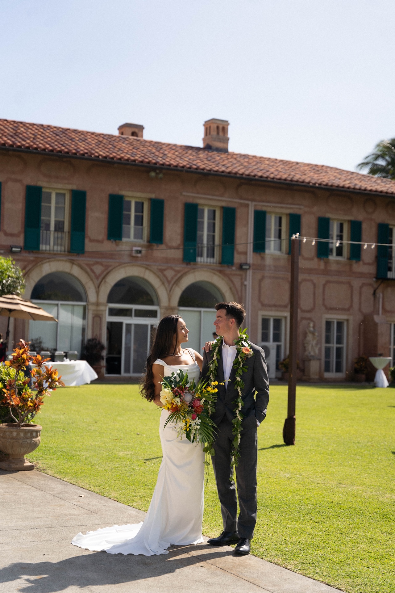 The couple stands on a lawn in front of an elegant estate, smiling at each other, the bride holding a colorful tropical bouquet.