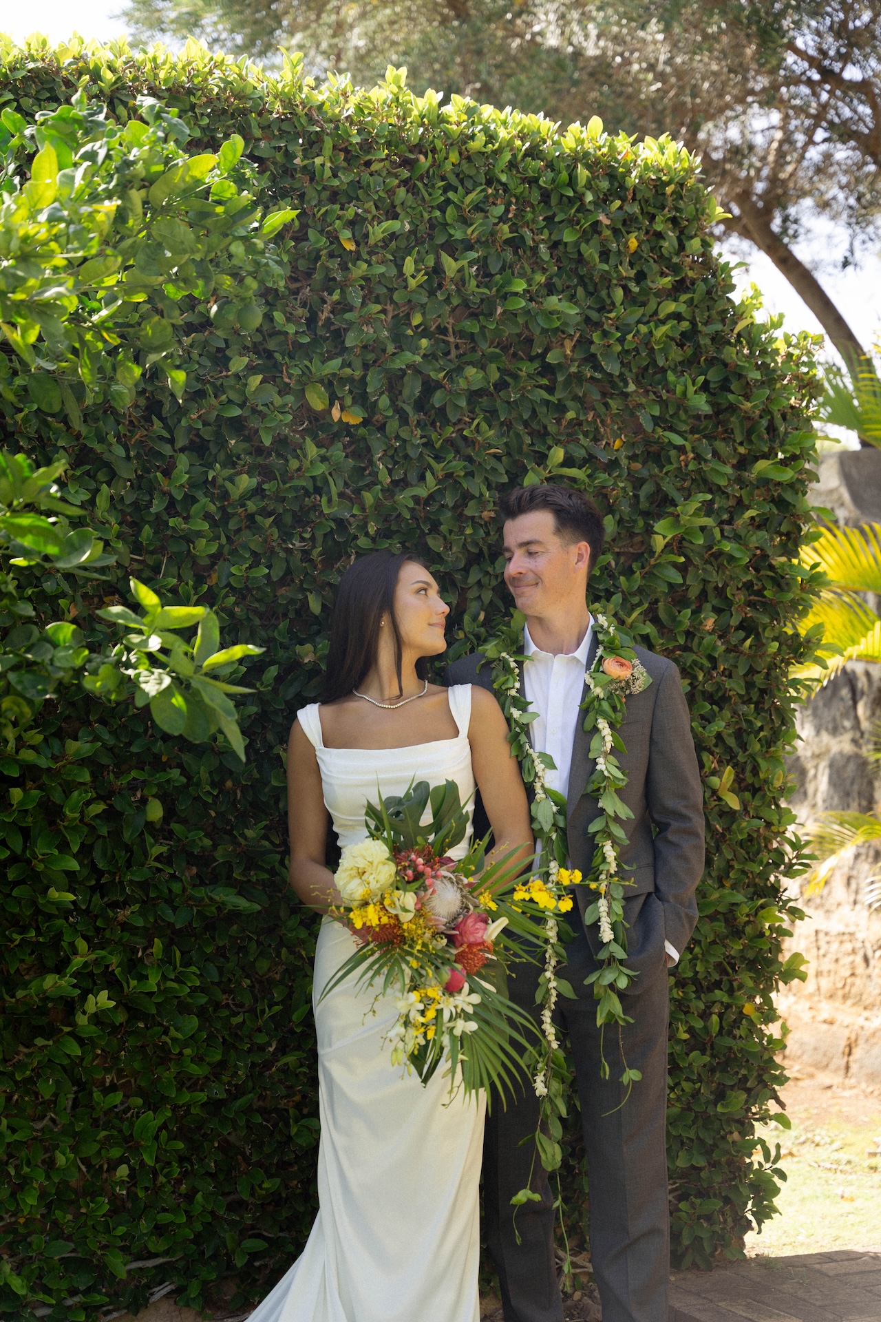 The bride and groom smile at each other while standing in front of a tall leafy hedge, her bouquet full of bright island florals.