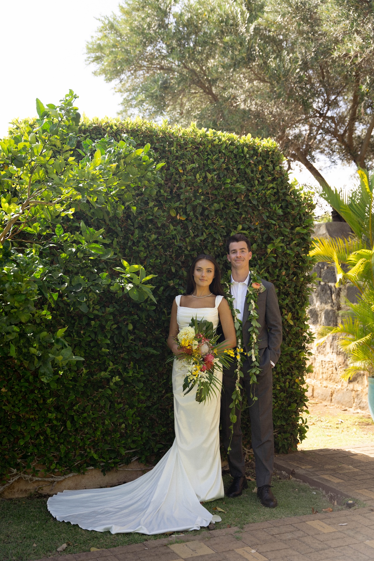 A formal portrait of the bride and groom standing against a manicured hedge, her train fanned out and bouquet in hand.
