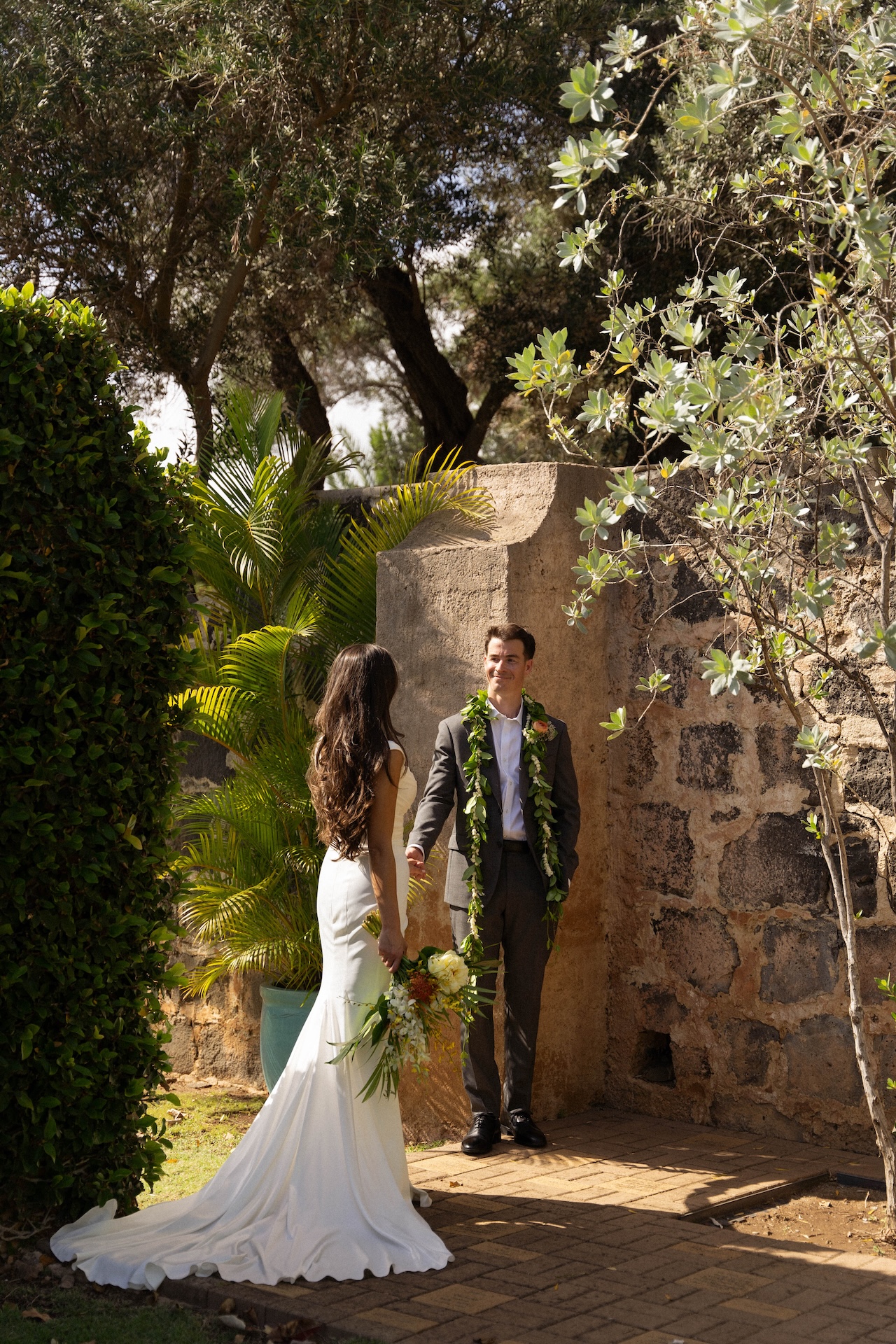 The groom reaches out to the bride as she walks toward him holding a tropical bouquet, framed by stone walls and palm trees — captured by a destination wedding photographer.