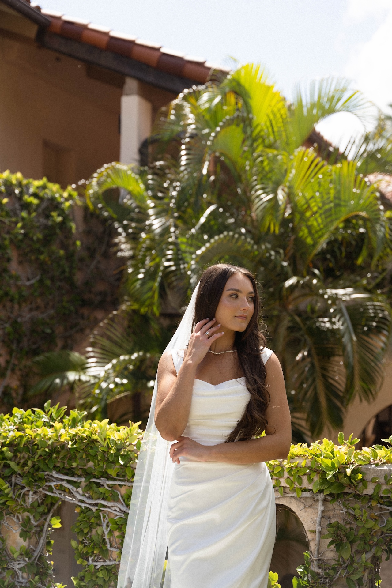 A close-up of the bride resting her hand on her shoulder, standing in front of palm trees and a weathered stone wall.