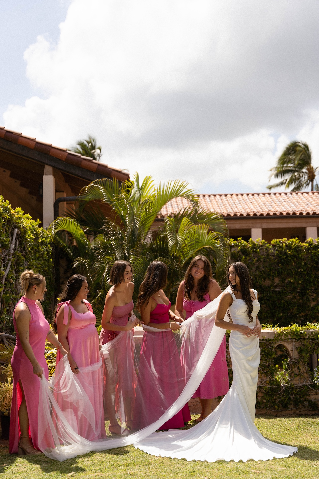 The bridal party surrounds the bride with laughter and movement as they help fluff her veil on the grass, mid-celebration.