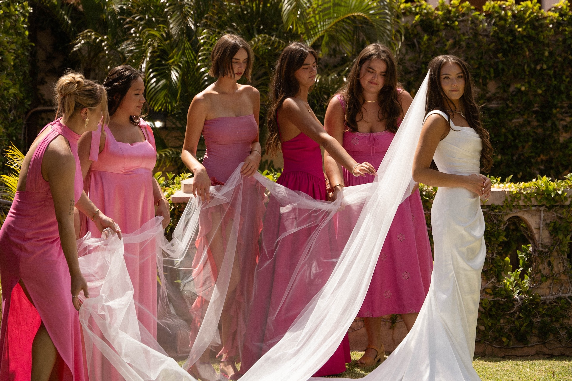 The bride stands in a lush courtyard surrounded by bridesmaids in pink dresses, who are helping arrange her long veil.