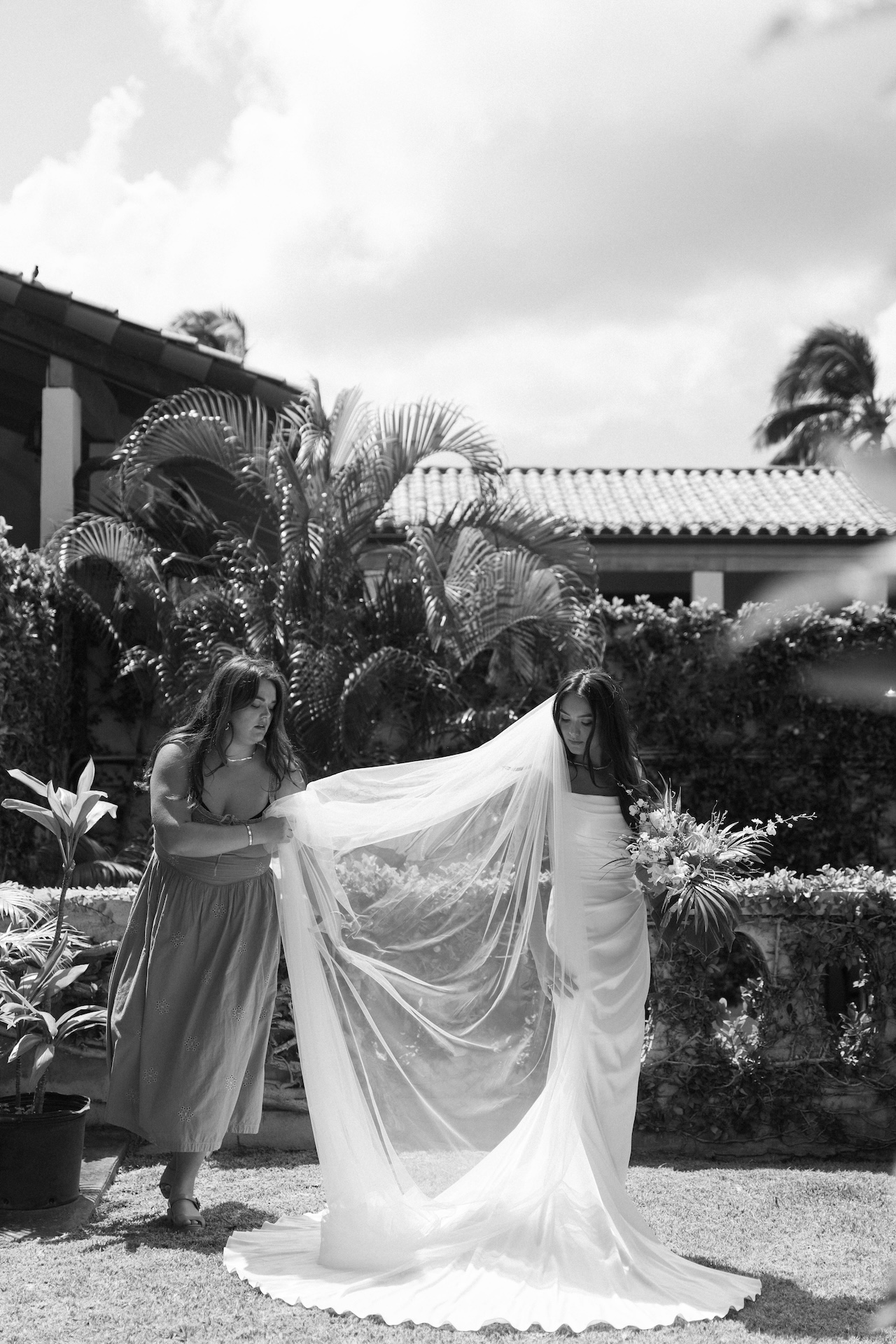 A bridesmaid gently adjusts the bride’s long veil in a garden courtyard, with lush palms and sunlight surrounding them — styled by a destination wedding photographer.