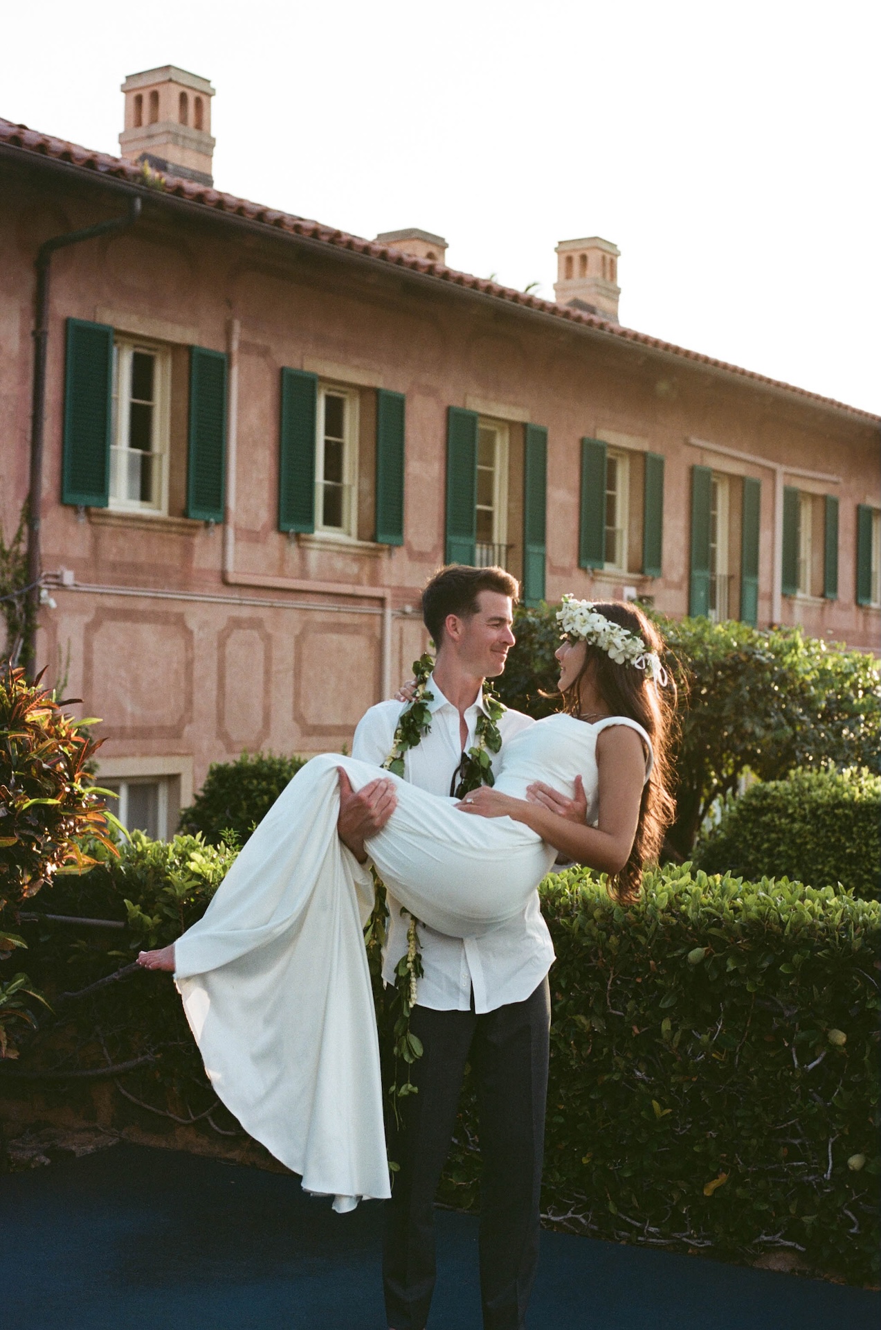 A groom carries his bride while smiling at each other in front of a vintage-style building with green shutters, captured by a destination wedding photographer.