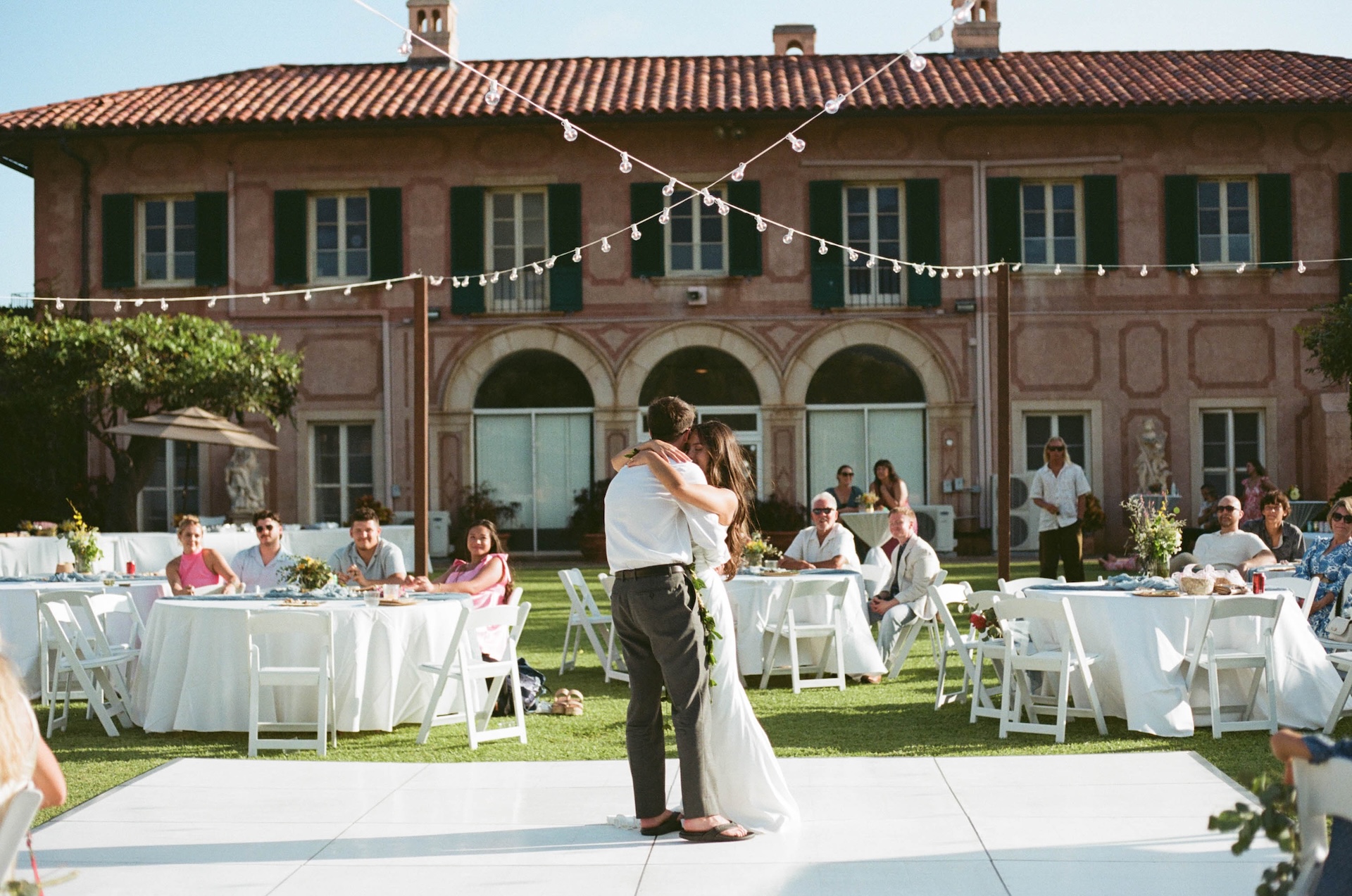The couple shares their first dance under twinkle lights on a lawn in front of a Tuscan-style estate, guests seated at round white tables.
